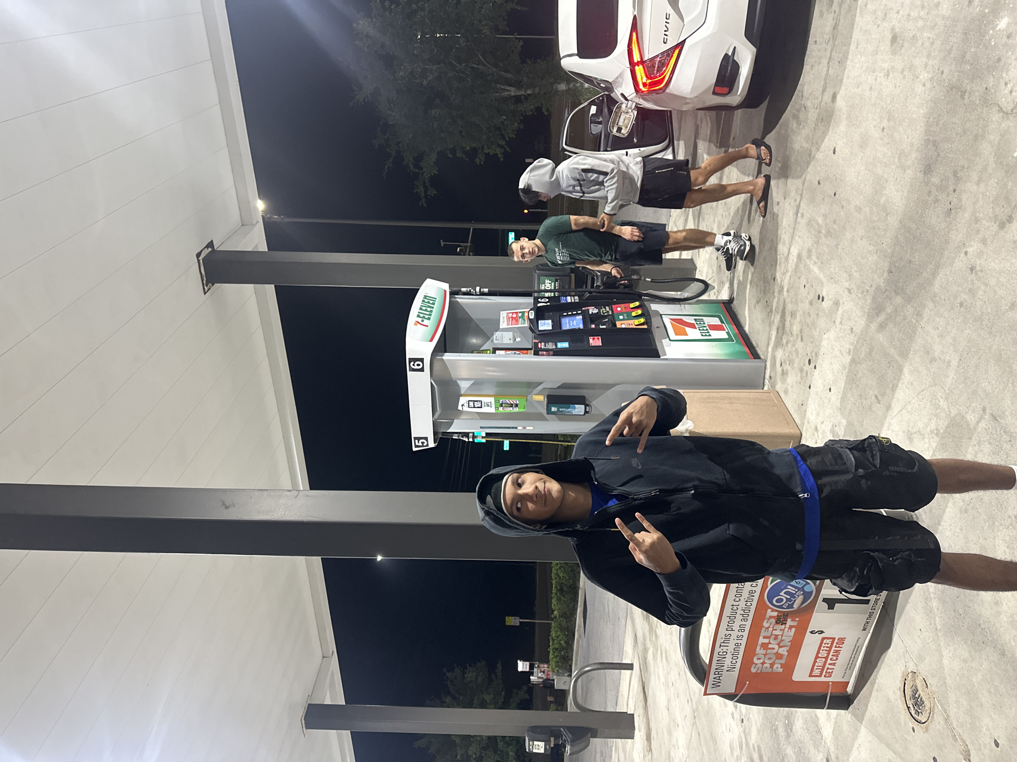 This nighttime scene shows a group of three individuals at a 7-Eleven gas station. A white Honda Civic is parked next to a gas pump, with the driver's side door open. One person, wearing a gray hoodie, is standing next to the car, while another in a green shirt is closer to the gas pump. The third person is near a display stand promoting "on!" nicotine pouches, holding his hands up, and his face is towards the camera. The environment is illuminated by the station's lighting. The location is Homosassa Springs, United States of America, based on the presence of a 7-Eleven gas station.
