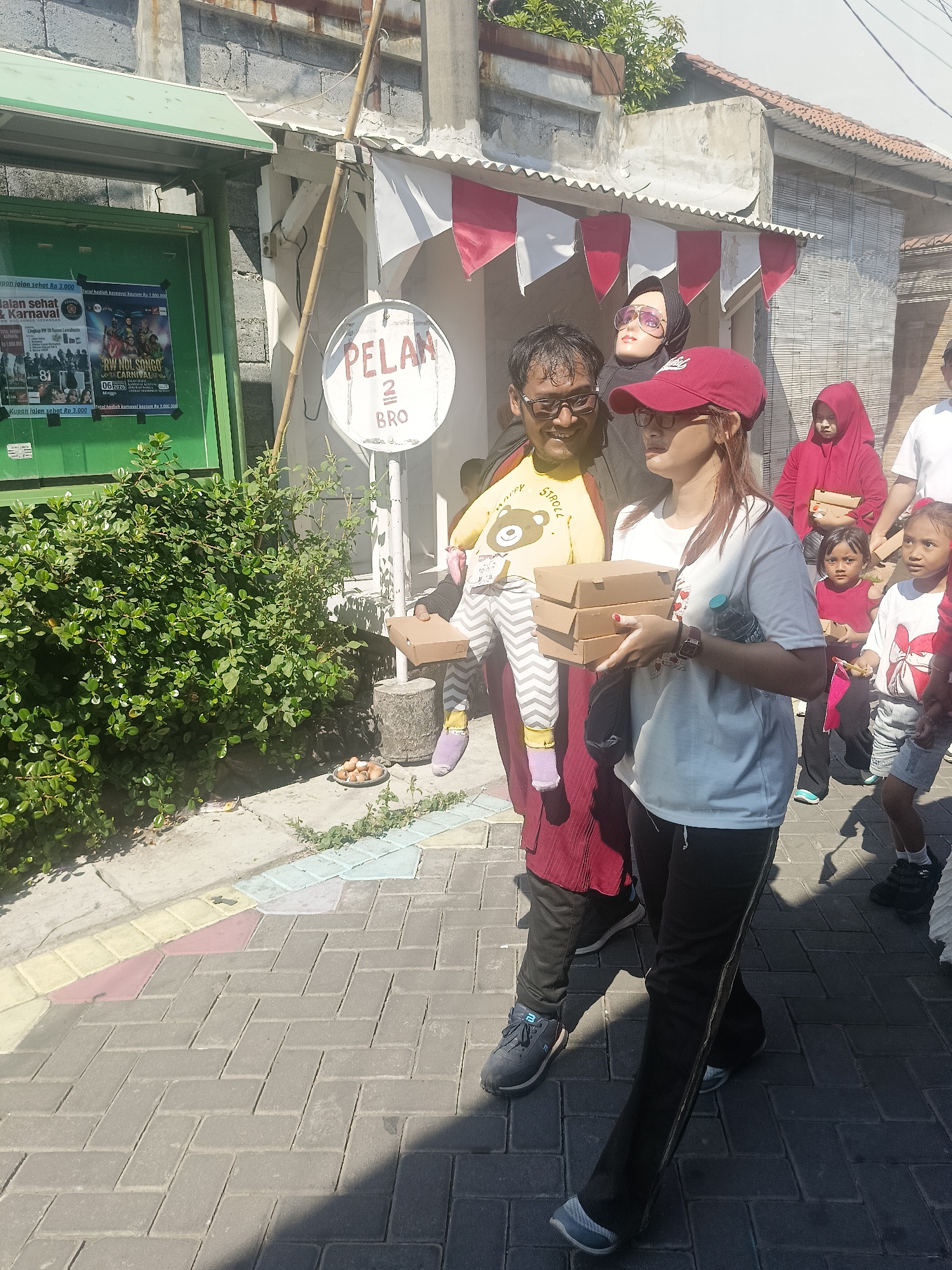 In the city of Gedangan, Indonesia, a group of people are walking on a paved street. The scene appears to be outdoors, with a building and a green structure on the left side of the image. Several individuals are visible, including adults and children. One man in the foreground is smiling and wearing glasses, holding a baby wearing a yellow shirt with a teddy bear design. Beside him, a woman in a red cap and white t-shirt is holding three brown cardboard boxes. Another woman in a red hijab and a red dress is also present, as are two young children, one in a red shirt and the other in a white t-shirt. The background shows a building with red and white banners and what looks like a sign with the text "PELAN 2 BRO". The street is paved with grey interlocking bricks, and there is greenery in the lower left corner. The lighting suggests it is daytime, with shadows cast on the pavement.