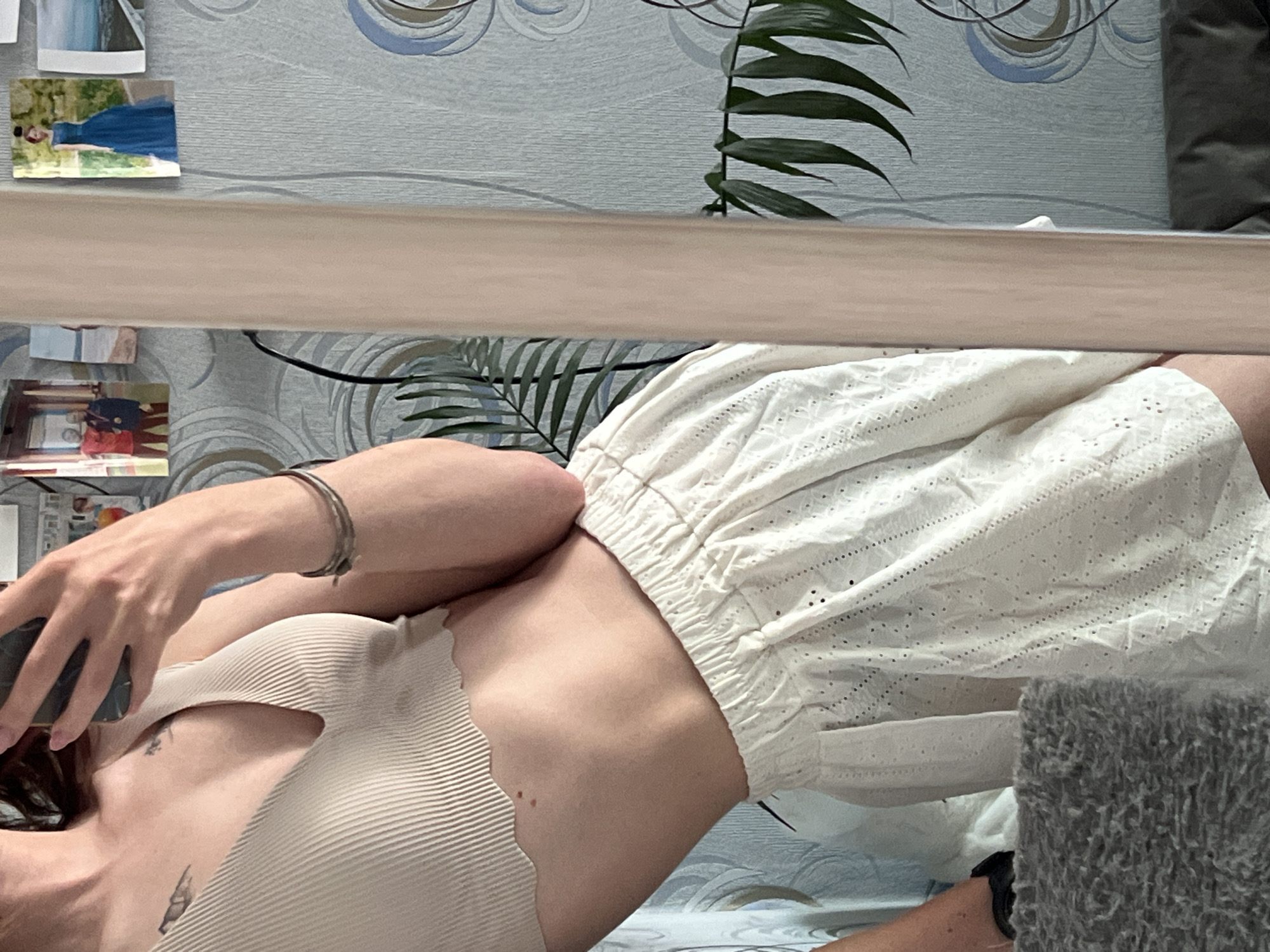 A person is lying down, wearing a light-colored ribbed top and a white, eyelet-style skirt. The person is holding a phone in their hand. The background includes a wall with a wallpaper featuring abstract patterns, some small framed pictures, and a potted plant. The setting appears to be indoors. It is not possible to discern if this image was taken in the city of Chaikovsky, Russia.