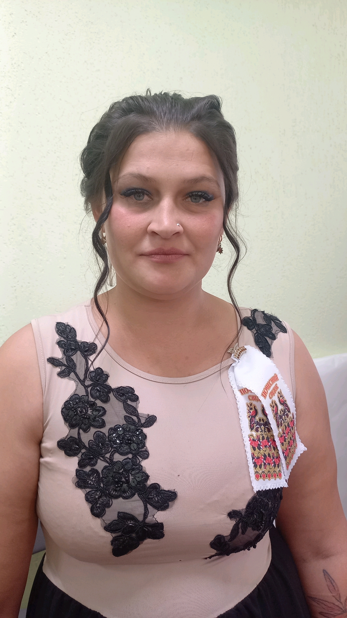 A medium shot photograph shows a woman with dark hair styled up and in loose curls around her face. She is wearing a light-colored sleeveless top with black lace floral appliques. A white ribbon with a colorful embroidered design, possibly a traditional motif, is pinned to her right shoulder. She is wearing earrings and a nose ring. The background is a plain, light-colored wall. The image appears to have been taken indoors. The woman is looking directly at the camera.