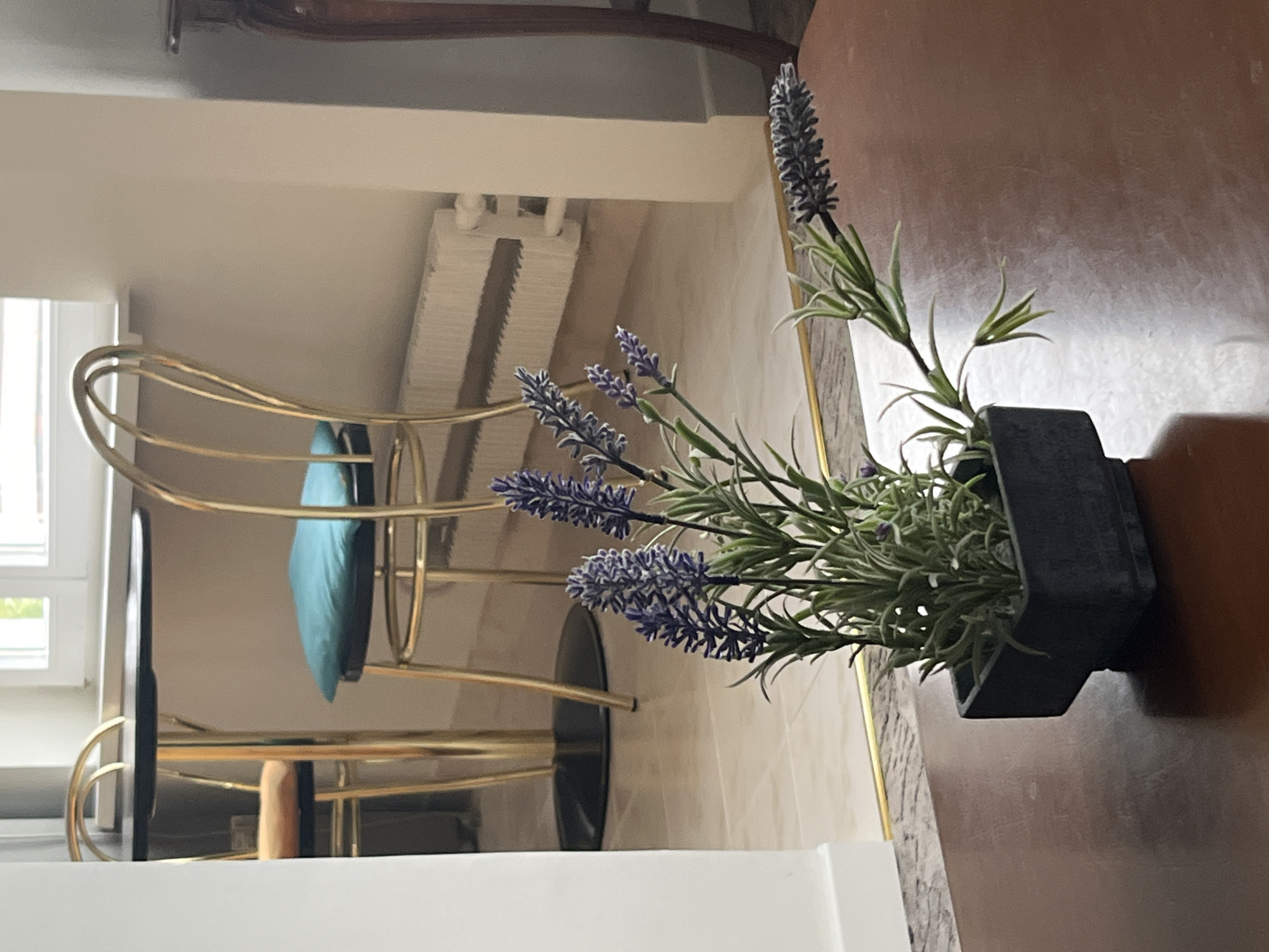 A faux lavender plant in a square dark grey pot sits on a brown wooden table. In the background is a gold metal chair with a blue cushion, a white radiator, and a window. The location is Moscow, Russia.