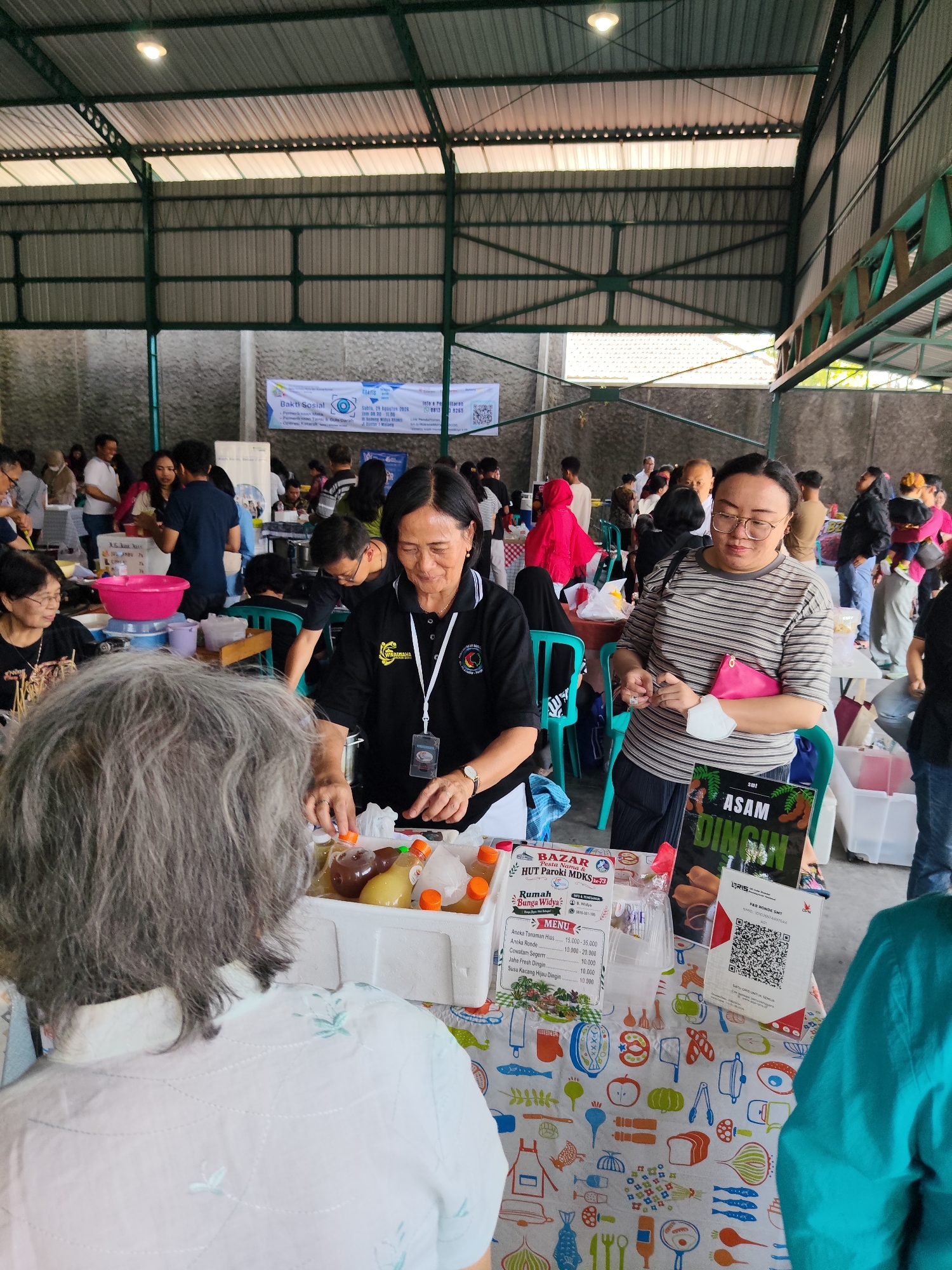 In a covered, open-air market in Malang, Indonesia, several people are gathered around food stalls. A woman in a black shirt with a white lanyard is serving drinks from a white cooler. To her right, a woman in a striped shirt holds a pink pouch and a mask. A table in the foreground displays bottles of drinks and a sign with the text "ASAM DINGIN". Other stalls and attendees are visible in the background, along with banners indicating a social event.