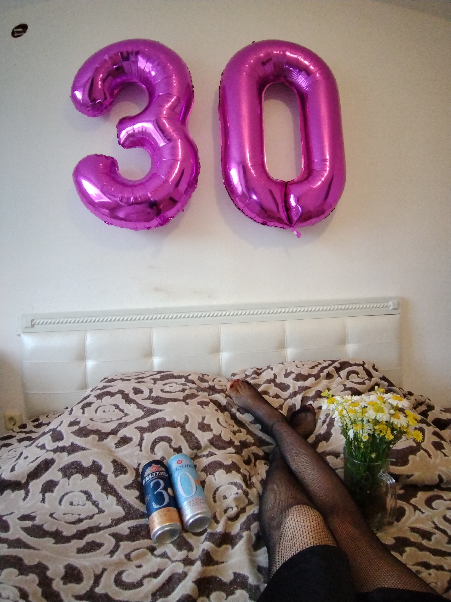 In a bedroom in Noviy, Russia, two large, shiny pink balloons in the shape of the numbers "3" and "0" are affixed to a white wall. Below the balloons, a white headboard of a bed is visible. The bed is covered with a comforter featuring a brown and beige ornate pattern. Two beverage cans, one blue and labeled "Baltika 3", and another light blue with white text, are placed on the comforter. To the right of the cans, a small bouquet of white and yellow daisies in a glass vase rests on the bed. A person's legs, wearing black fishnet stockings and red nail polish on their toes, are crossed on the comforter. The person is wearing a black garment. The overall scene suggests a birthday celebration, possibly for someone turning thirty.