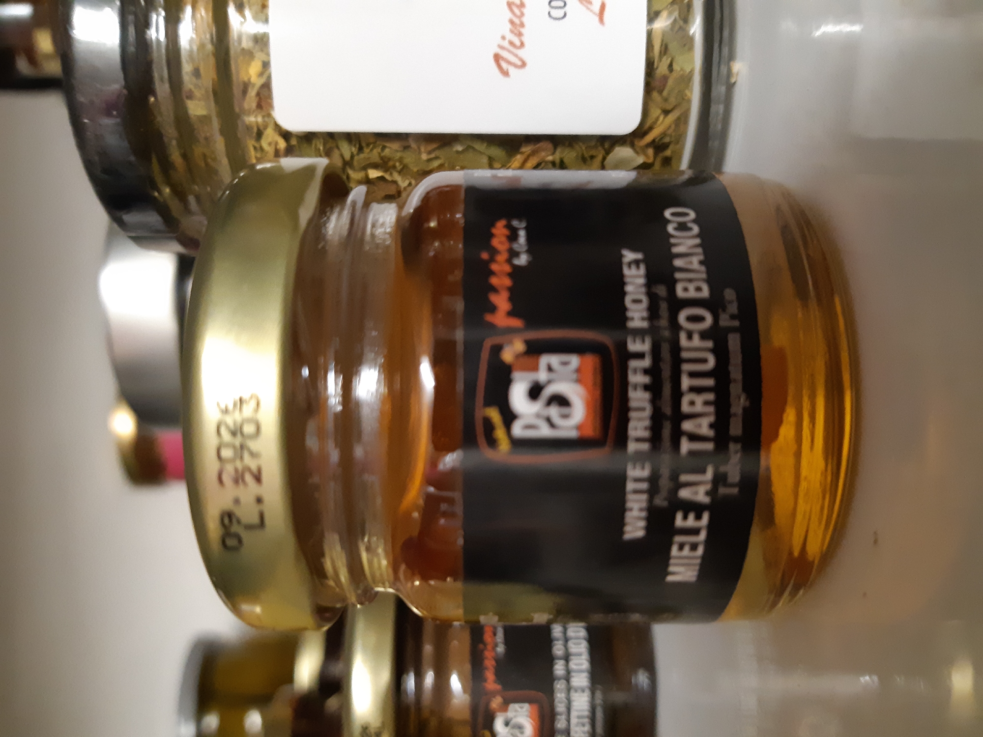 The image displays a close-up of three glass jars positioned on a light, reflective surface, likely a shelf. The central and most in-focus object is a small, clear glass jar containing a golden-amber liquid, identified by its black label as "WHITE TRUFFLE HONEY" / "MIELE AL TARTUFO BIANCO" by "P.STA passion by Cova C," derived from "Tuber magnatum Pico." This jar has a gold-colored metallic screw-on lid, which is stamped with "09.2026" and "L.2703."
To the left and slightly behind the central jar, another blurred glass jar with a gold lid is visible. Its partial label indicates "passion" and phrases like "SLICES IN OLIV" and "PETTINE IN OLIO DI," suggesting a product containing truffle slices in olive oil. To the right and also blurred, a wider glass jar with a silver-colored metallic lid contains dried green herbs or spices, possibly oregano. A partially visible white label on this jar shows text including "Vina" and "CO." The background behind the jars is a uniformly light, out-of-focus surface. No individuals, direct actions, or interactions are depicted. The exact location within the city of Plovdiv, Bulgaria, cannot be determined from the visual evidence.