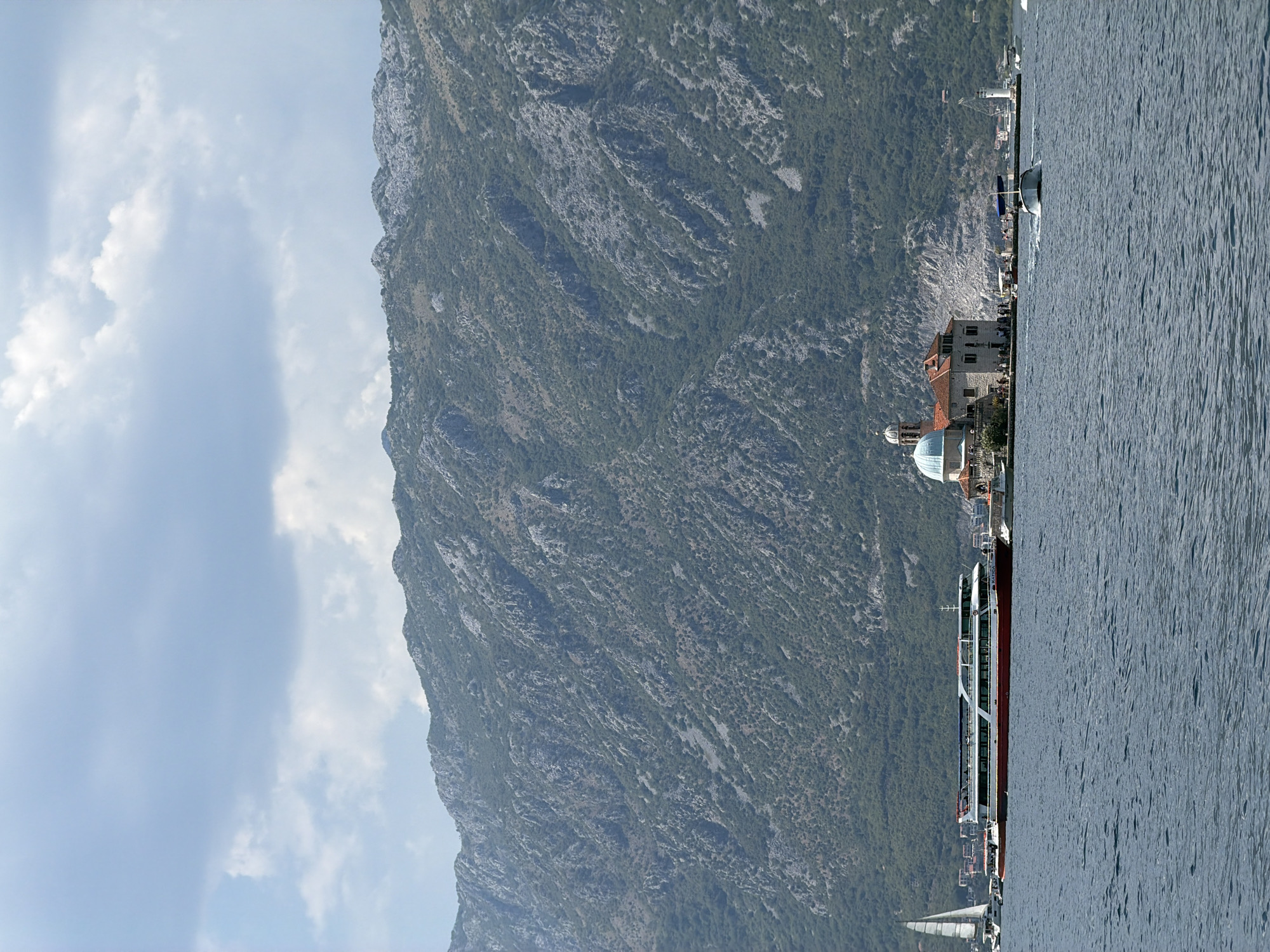 The image depicts a view of the Bay of Kotor, near the town of Perast, Montenegro. In the foreground, a body of water is visible, with ripples on its surface. Several boats are present on the water. To the left, a white sailboat with its sail up is partially visible. Dominating the center-left of the image is a large, multi-deck passenger ferry, predominantly white with a red hull and a blue stripe along its upper deck. The ferry appears to be docked or moving slowly. To the right of the ferry, an island features a building complex with a prominent blue-domed church and red-tiled roofs. Several people are standing on the island near the buildings. A white lighthouse is also situated on the right side of the island. In the middle ground, a small motorboat is creating a wake as it moves through the water. The background is dominated by a steep, mountainous landscape covered with dense green vegetation and rocky outcrops. The sky above is partly cloudy, with large, soft white and gray clouds against a pale blue backdrop. The overall scene conveys a sense of a tranquil coastal environment with a mix of natural beauty and human activity.
