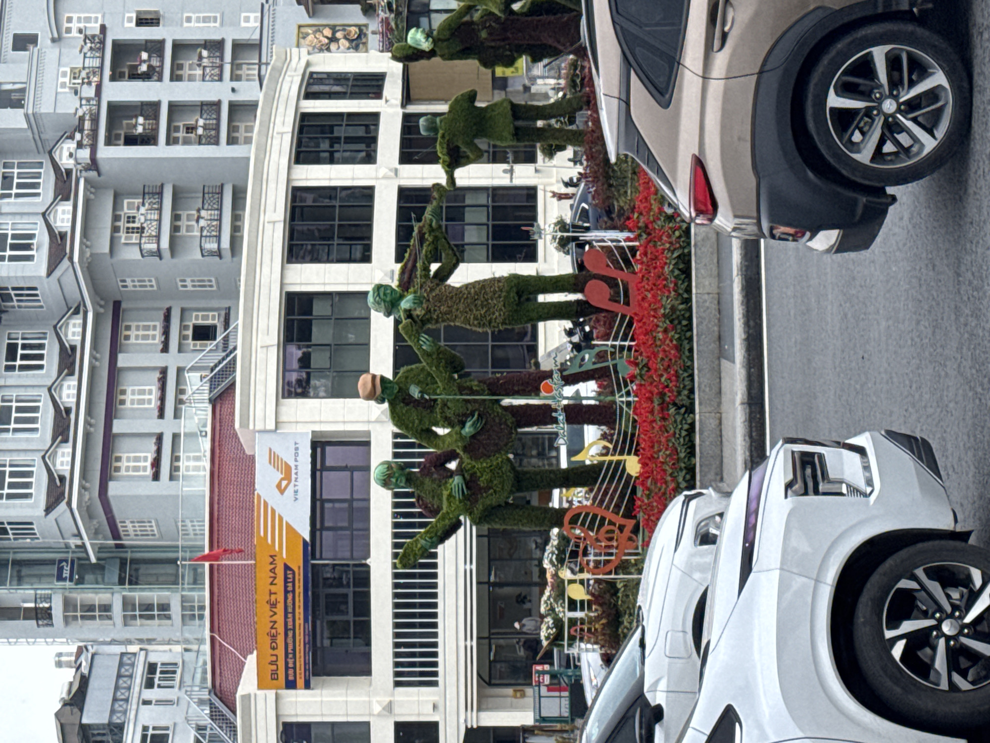 This daytime image, captured in Da Lat, Vietnam, displays a vibrant street scene with architectural and decorative elements. On the left side of the frame, a multi-story building with numerous windows and balconies dominates, featuring a red-tiled roof section and modern gray facades.
In front of this building, a creative display of topiary figures resembling musicians is arranged along a sidewalk or planter bed. These green, human-shaped topiaries are depicted playing various instruments, including violins and a double bass, with some musical notes as decorative accents nearby. The figures are surrounded by a long planter overflowing with bright red flowers, adding a splash of color to the scene. Part of a sign or decoration reads "Dalalato Garden."
To the right of the topiary display, two cars are prominently parked on what appears to be a street or parking area. The closest car to the viewer is a white SUV, with another beige SUV visible behind it.
A notable feature is a tall sign attached to the building on the left, displaying "VIETNAM POST" and "BƯU ĐIỆN VIỆT NAM." Below this in smaller text, "BƯU ĐIỆN PHƯỜNG XUÂN HƯƠNG, ĐÀ LẠT" is visible, directly confirming the location within Da Lat. A small red flag is also affixed to the signpost. The weather appears clear, suggesting a sunny or bright day.