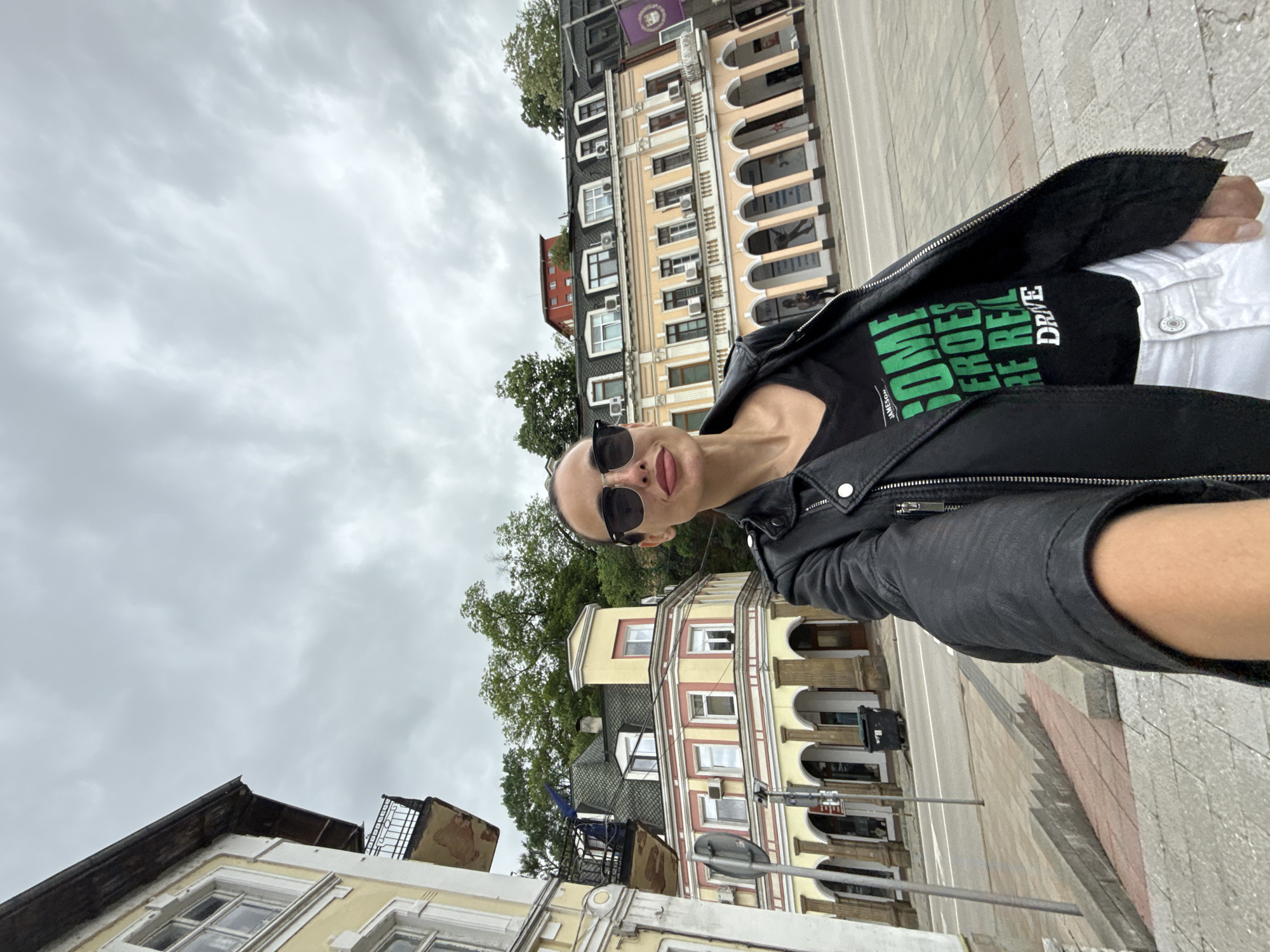 A woman wearing sunglasses and a black leather jacket stands outdoors on a city street. She is wearing a black t-shirt with green writing and a white shirt underneath. Buildings stand in the background, with arched windows and various colors, including beige, yellow, and red. Trees peek out from behind the buildings, and the sky is overcast with gray clouds. The location is Lovech, Bulgaria. The woman is taking a selfie.