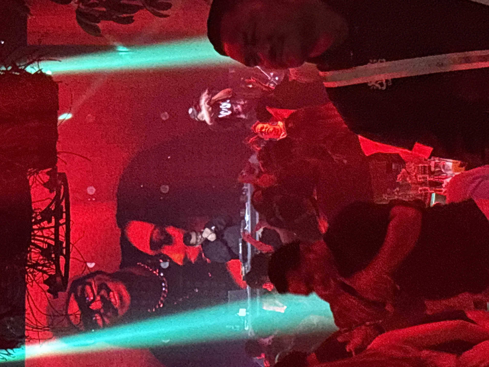 The image depicts a dimly lit indoor nightclub scene in North Jakarta, Indonesia, characterized by pervasive red and green lighting. In the foreground, a man is visible from the chest up, facing to the right, with his profile partially obscured by darkness. He wears a black shirt and appears to be looking towards the right side of the frame. In the mid-ground, a DJ is positioned behind a console, wearing a black long-sleeved shirt and glasses, with his hands near the equipment. A crowd of people is gathered in front of the DJ, some with their hands raised. The background features large screens displaying abstract red patterns, and decorative elements hanging from the ceiling. A vibrant green light beam cuts across the scene from the left. The overall atmosphere suggests a lively event or party.