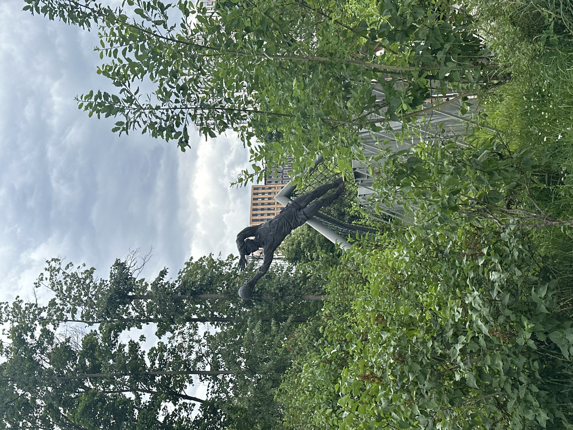A dark bronze sculpture of a soccer goalkeeper is positioned in an outdoor setting, appearing to catch a ball. The sculpture is centrally located, partially obscured by lush green foliage and trees in the foreground. The goalkeeper is in a dynamic pose, leaning forward with arms outstretched. Behind the sculpture, a modern building with a light-colored facade and rectangular windows can be seen through a gap in the trees. The sky is overcast with gray clouds. The surrounding environment suggests a park or public space with a blend of natural elements and urban architecture.