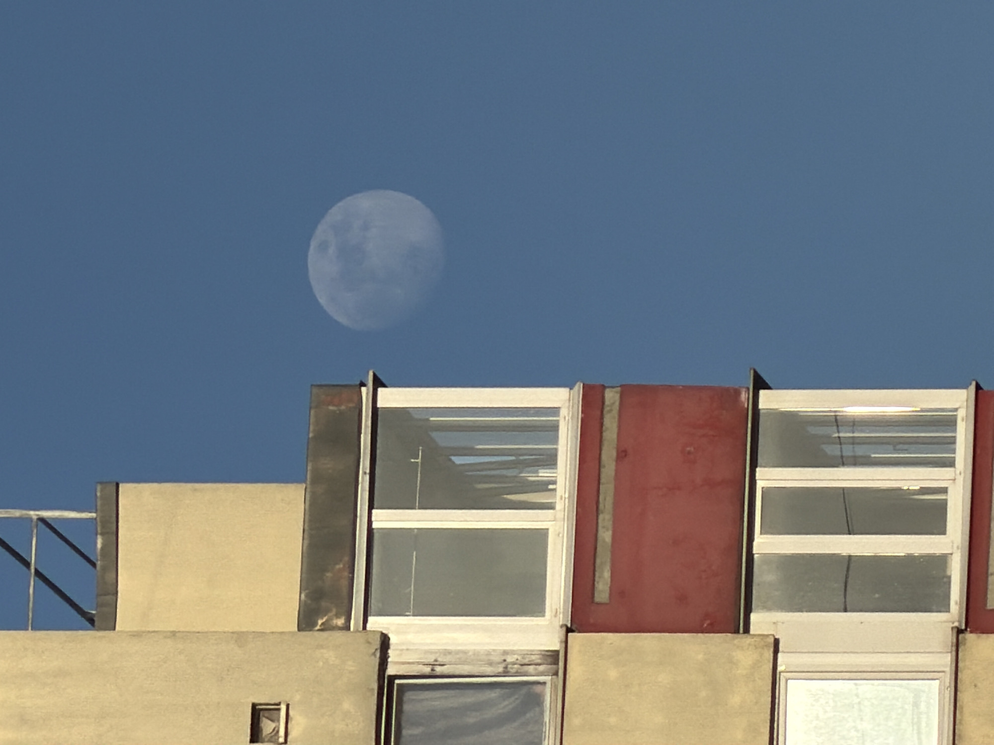The image shows a partial view of a building in daylight, with the moon visible in the clear blue sky above. The building appears to be multi-storied and features various rectangular window frames. The building's facade consists of sections in colors like beige, red, and dark gray. There is a metal railing visible at the top left. The windows have white frames and some show reflections. There are no people visible in the image. The location is purportedly the city of Chaikovsky, Russia, though no specific identifiers of the city are evident.