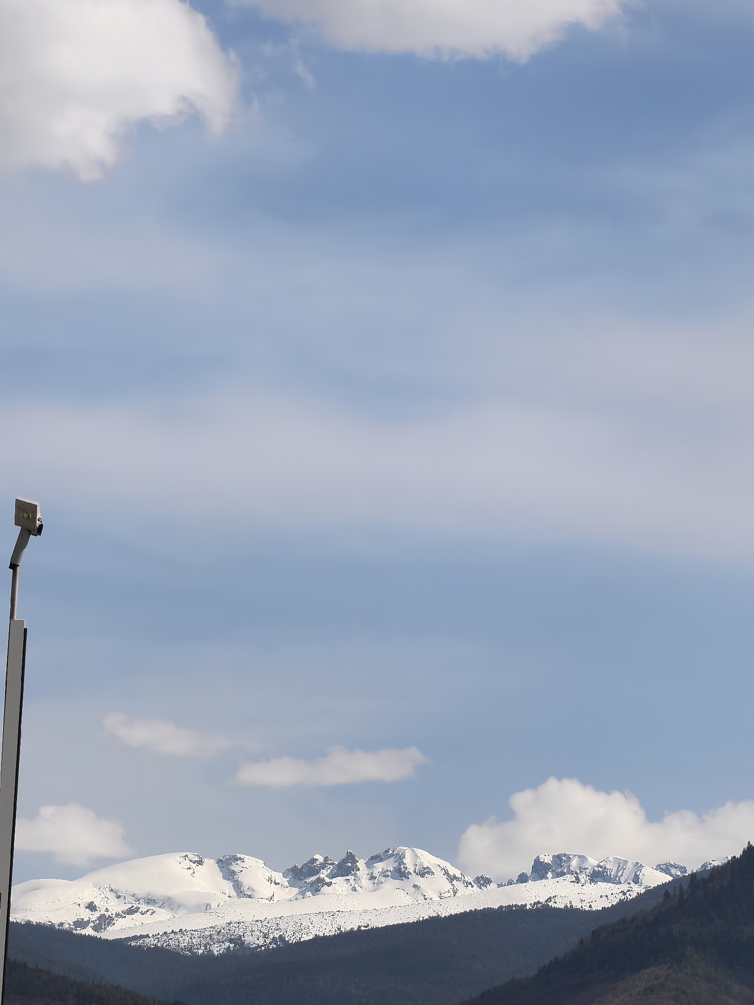 The image displays a vast mountain landscape under a clear blue sky dotted with scattered white cumulus clouds. The sky occupies the majority of the frame, particularly the upper two-thirds.
In the mid-ground, a prominent range of snow-capped mountains stretches horizontally across the scene. Their rugged peaks and upper slopes are covered in pristine white snow, contrasting with some visible darker rock formations. Below these snowy peaks, the foreground is dominated by darker, dense coniferous forests that cover the lower mountain slopes and hills, suggesting a cooler climate.
On the far left side of the image, a tall, light-colored pole or mast is partially visible, extending upwards from the bottom of the frame. Attached to its top is a small, rectangular, light-gray device, possibly a sensor, camera, or part of a weather station, featuring a small dark, circular element on its side.
The scene is captured during the daytime, likely late morning or early afternoon, given the bright illumination and distinct shadows. The weather appears fair and calm, with a gentle, diffused light. No people are visible, and there is no discernible text present on any of the objects or within the landscape. The overall atmosphere conveys a sense of natural beauty and tranquility in a mountainous environment.