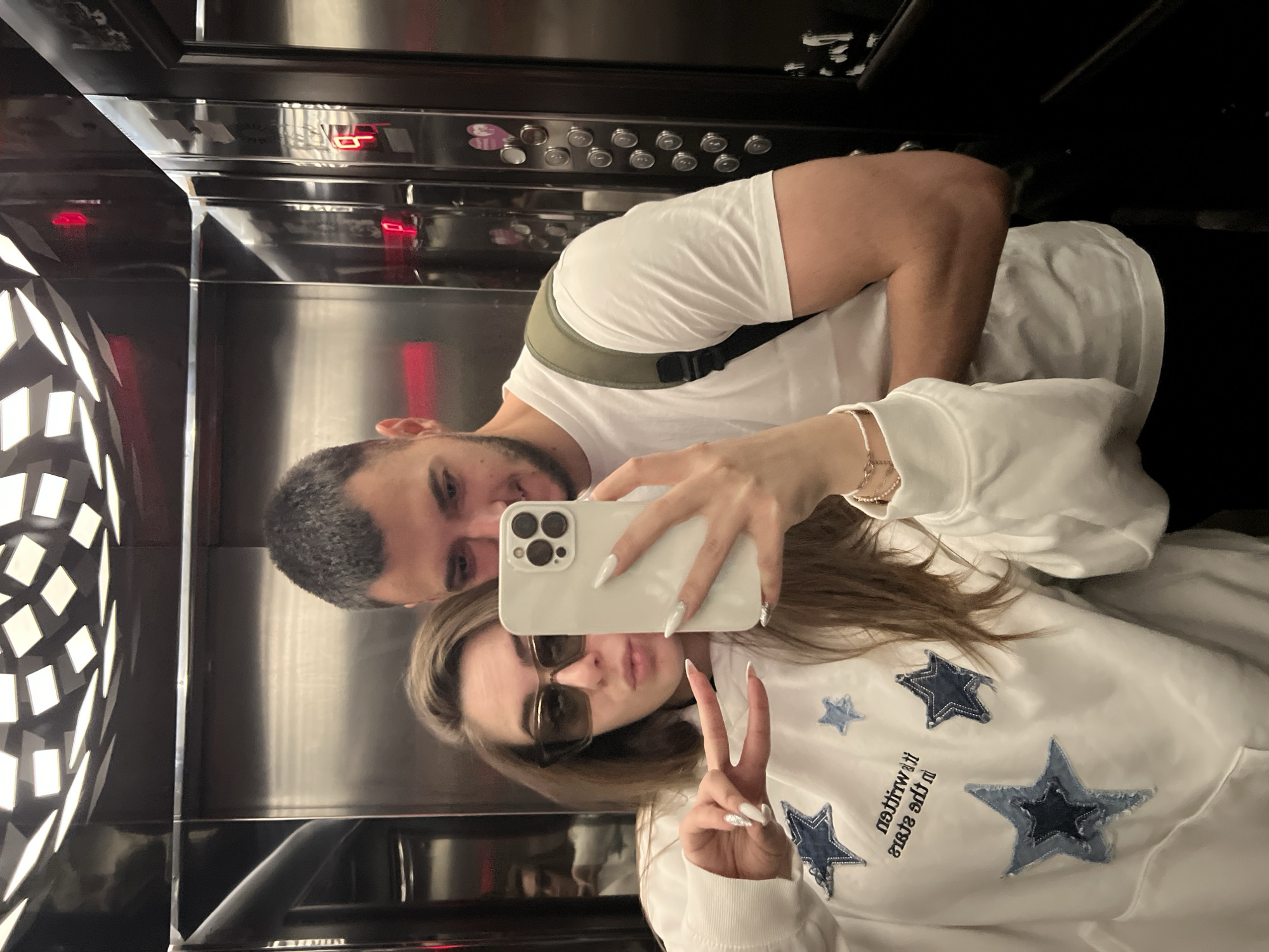 A rotated, vertical mirror selfie captures a man and a woman inside a modern elevator in Plovdiv, Bulgaria. The woman, positioned on the right side of the frame, is taking the photo with a white smartphone. She wears dark sunglasses, a white hooded sweatshirt with multiple blue denim star patches, and a bracelet on her right wrist. Her left hand is raised in a peace sign, displaying long, white fingernails. Reflected and mirrored text on her sweatshirt is partially visible.
Behind and to her left is a man with a beard, wearing a white t-shirt and an olive-green or dark gray strap, possibly from a bag. He looks towards the camera.
The elevator interior features polished metal walls, creating significant reflections of the subjects and the internal lighting. In the background, an elevator control panel with multiple silver buttons is visible through reflections. A red LED display clearly shows the number "6," indicating the current floor. To the left, a decorative panel with a geometric pattern is reflected in the elevator's surfaces. The lighting is artificial and consistent with an indoor environment.