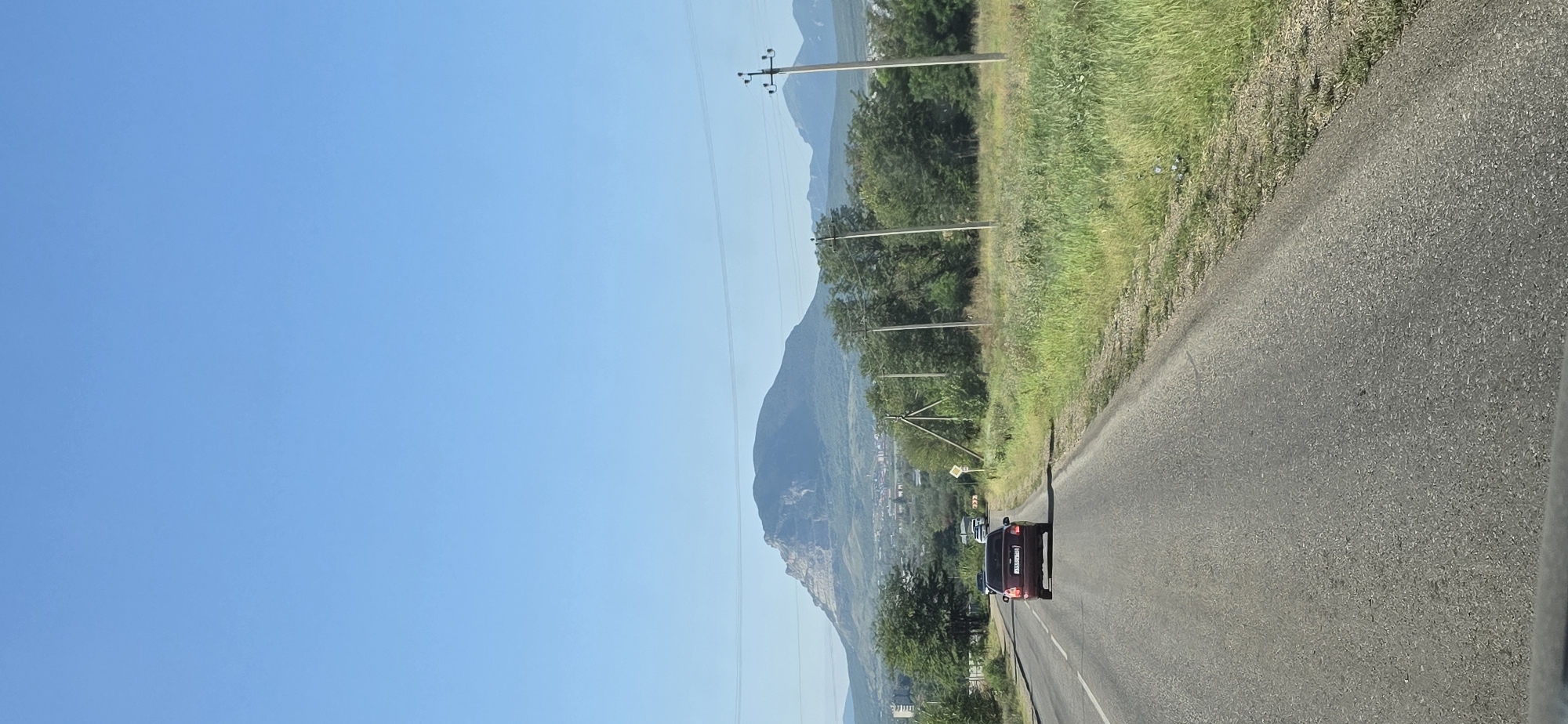 The image shows a view from a vehicle traveling on a paved road with a single lane in each direction. A dark red car is driving ahead on the same lane. To the right of the road, there is a grassy area with scattered bushes and trees, and several utility poles. In the distance, a large, rounded mountain with a lighter colored, rocky summit is visible under a clear blue sky. The area surrounding the mountain appears to be a valley with some buildings and trees. The overall scene suggests a rural or semi-rural setting.