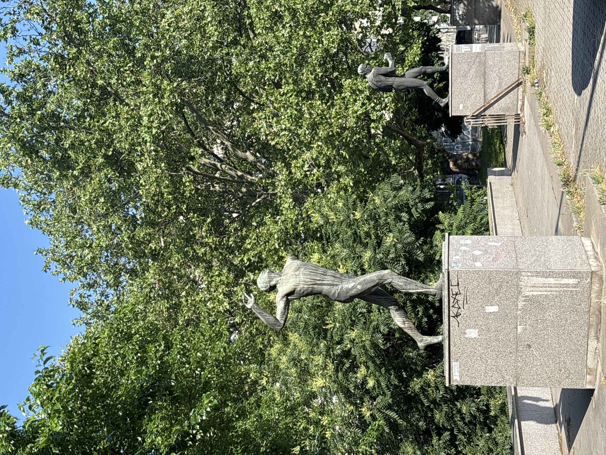 Two bronze statues of male athletes stand on granite pedestals in a park-like setting with lush green trees and a bright blue sky. The statue in the foreground depicts a lean figure in a dynamic pose, with its left arm raised and head tilted back, appearing to be mid-action, possibly throwing a discus or javelin. The statue in the background is smaller and further away, also appearing to be in a running or throwing posture. The pedestals are weathered, with one in the foreground showing graffiti and water stains. The ground is paved with cobblestones. The image appears to be taken in the South Park in Sofia, Bulgaria, a location known for its sculptures and green spaces.