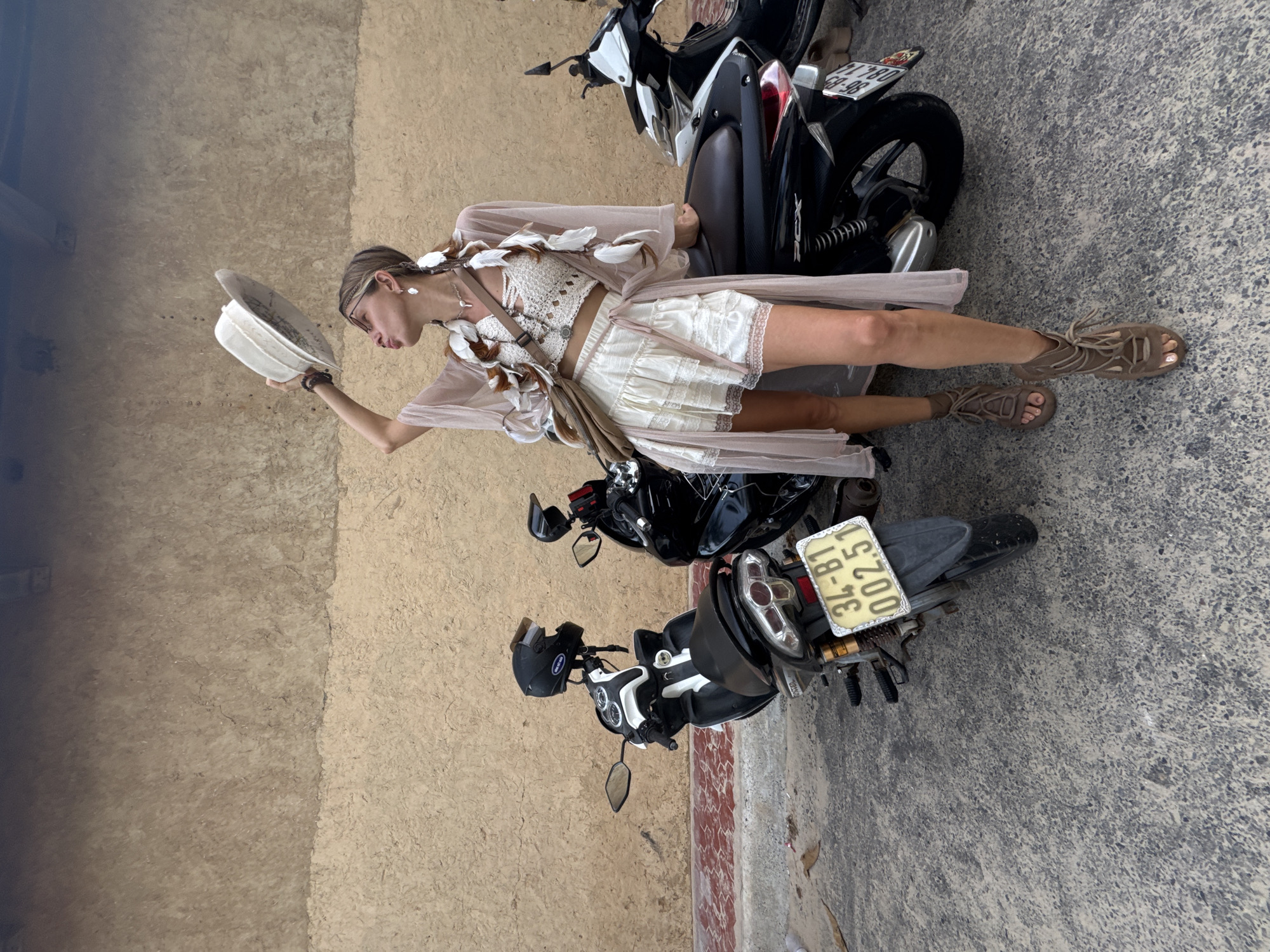 A woman stands next to two parked motorcycles in Mũi Né, Vietnam. She is wearing a light-colored, bohemian-style outfit consisting of a lacy white crop top, a white ruffled skirt, and a sheer, flowy kimono. She has a wide-brimmed hat in her right hand, held above her head, and is puckering her lips as if about to kiss it. Her hair is styled in braids adorned with feathers and a headband. She is also wearing gladiator-style sandals and a crossbody bag. The motorcycles appear to be common commuter models. The background consists of a textured, earthy-toned wall. The ground is paved with asphalt and gravel. The scene suggests a casual, perhaps artistic or adventurous, setting within the city.