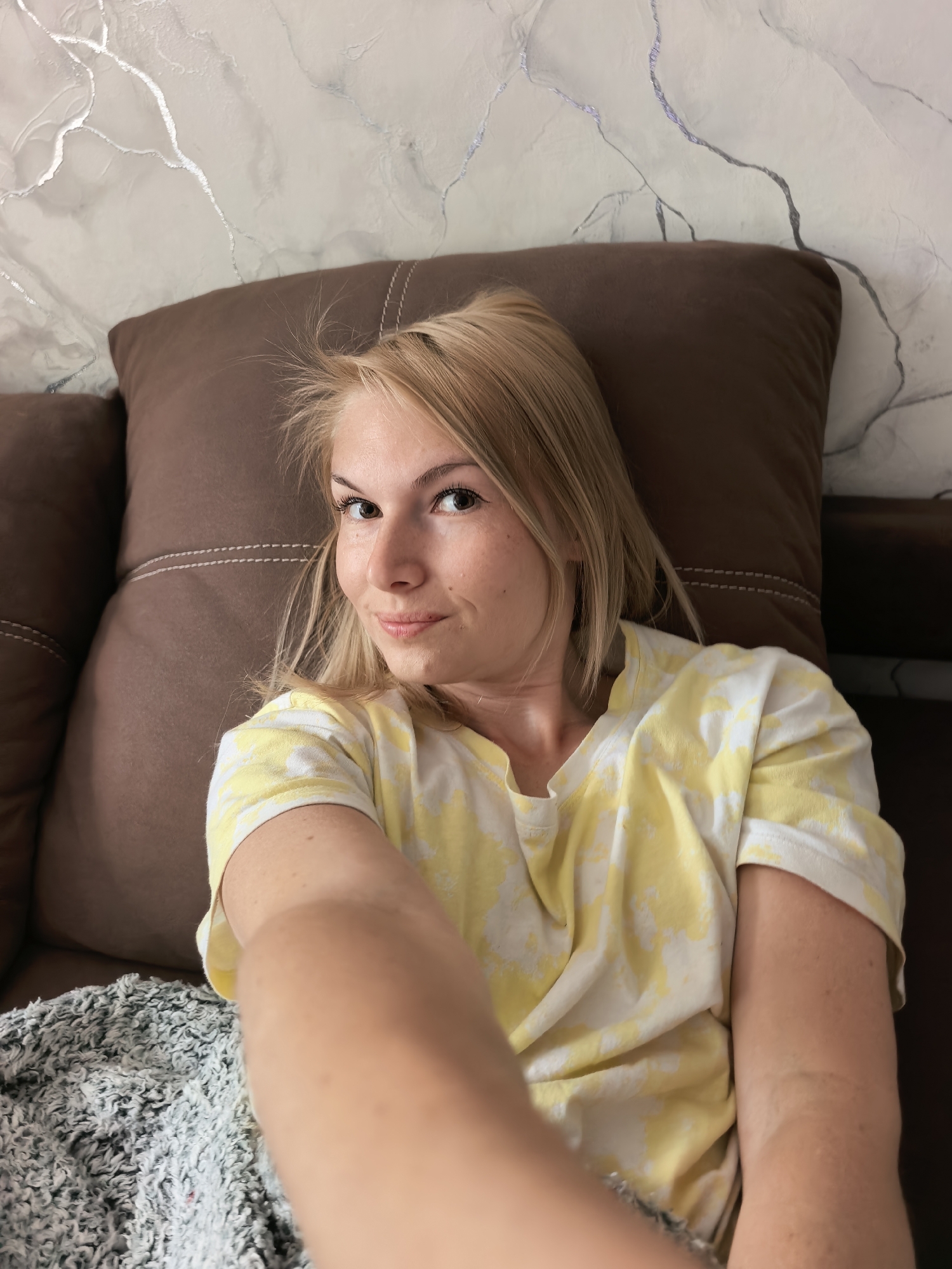 A fair-skinned woman with blonde hair is taking a selfie while lying on a brown couch with a brown pillow behind her head. She's wearing a white t-shirt with yellow patterns. Part of a grey and white blanket is visible on the couch. The background shows a wall with a white and grey marble-like pattern. The location is unknown and cannot be determined to be the city of Sterlitamak, Russia, based on the provided information.