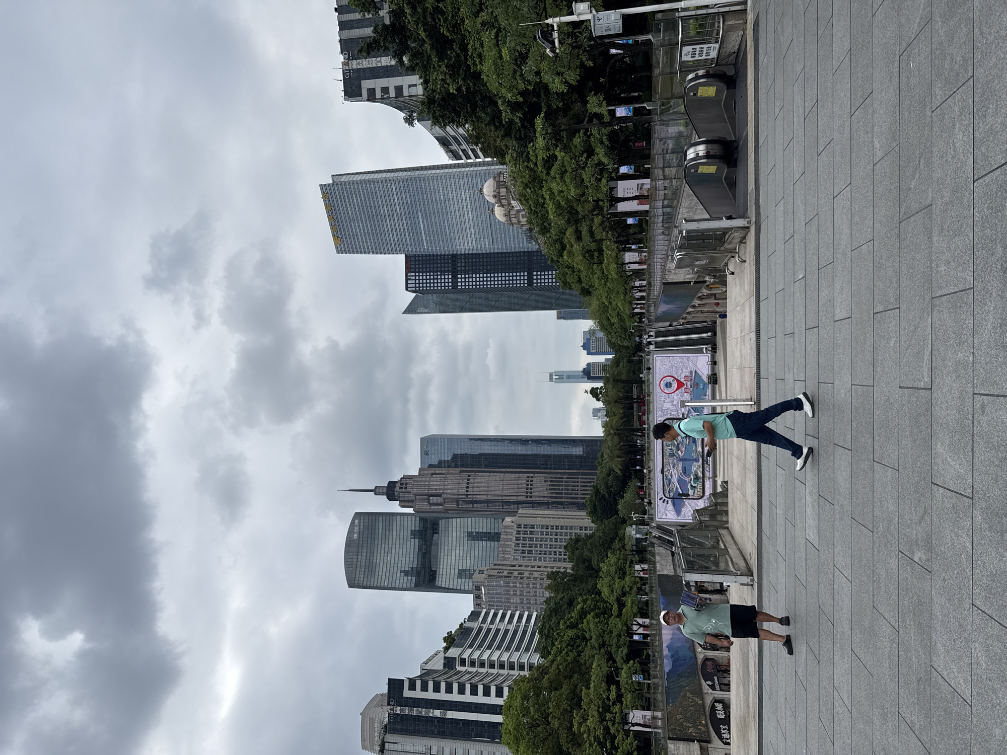 The image depicts an outdoor urban scene in Tianhe District, China, under a cloudy sky. In the foreground, two men are walking across a paved area. One man, wearing a light green polo shirt and dark shorts, is stationary and holding a tablet. The other man, in a turquoise t-shirt and dark pants, is walking towards the right. Behind them, a plaza area features escalators leading down, metal barriers, and banners. Beyond this, a lush green park area separates the plaza from a cluster of modern skyscrapers. The architecture of the buildings varies, with some featuring glass facades and others exhibiting more traditional design elements. The environment appears to be a public space within a densely developed urban district.
