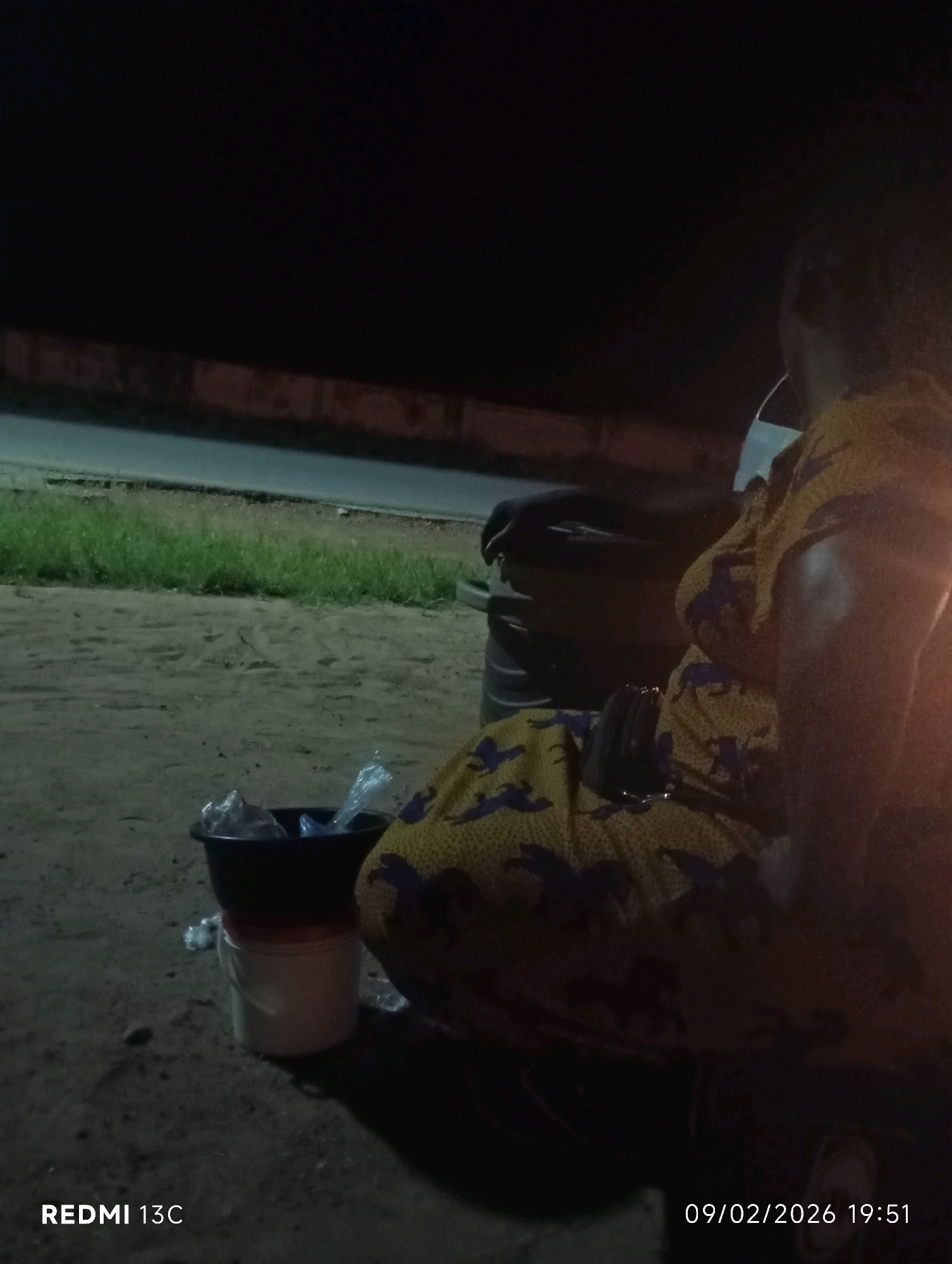 The image shows a woman sitting on what appears to be a motorcycle at night, in Shendam, Nigeria. She is wearing a yellow and purple patterned dress. In front of her, there are two containers: a black bowl on top of a white bucket. The background shows a grassy area next to a road, with dark, dimly lit surroundings. The bottom left corner of the image displays the text "REDMI 13C", and the bottom right corner shows "09/02/2026 19:51", indicating the date and time the photo was taken.