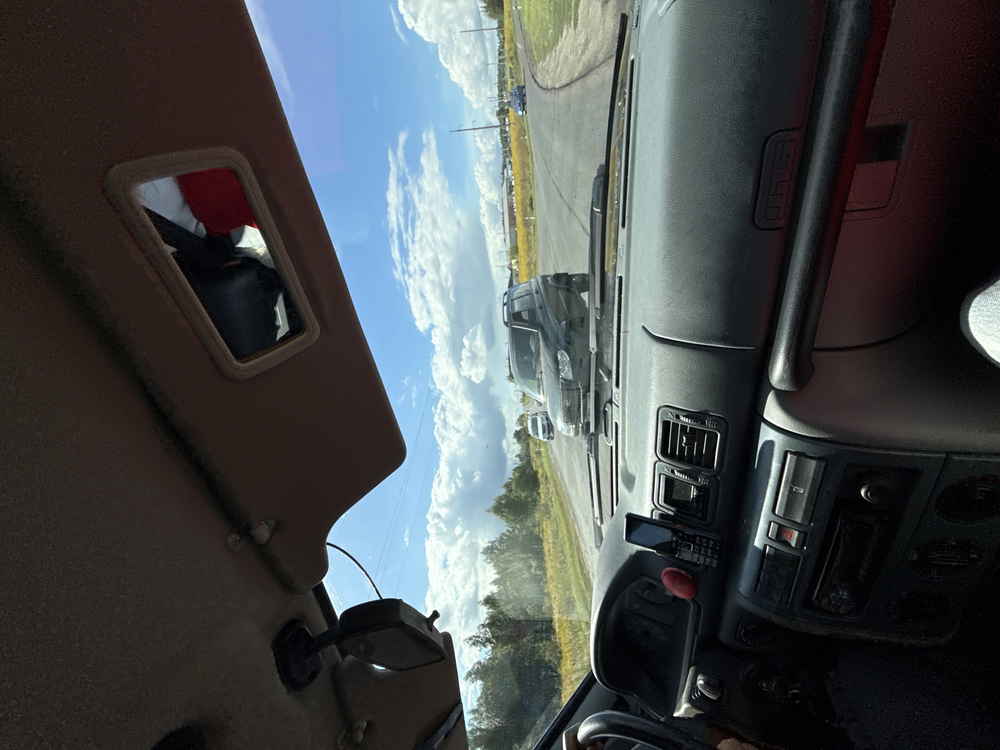 The image shows the interior of a car, looking out through the windshield at a road and its surroundings. The dashboard is visible in the foreground, with a cell phone resting on top of it. A car is driving on the road, and there are trees and fields in the background. The sky is blue with some clouds. The location is specified as Razvetye, Russia.