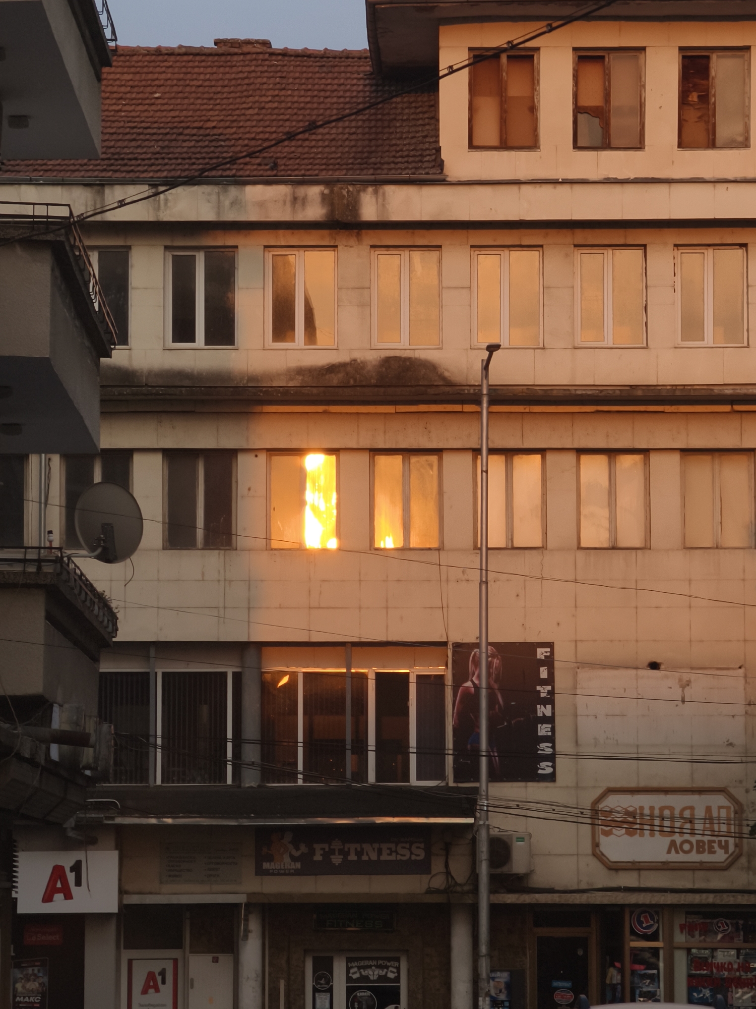 The image depicts a multi-story building in the city of Lovech, Bulgaria. The building has a weathered, beige facade with numerous windows. The upper stories feature rows of windows reflecting the warm tones of either sunset or sunrise, with some windows showing clear reflections of the sky and others appearing opaque and tinted orange. The roof is tiled in a reddish-brown color.
In the mid-section of the building, one window exhibits a particularly bright, fiery reflection, suggesting direct sunlight. A streetlamp stands in front of this section, obscuring part of the facade. Below the windows, advertisements and signs are visible. One prominent sign displays the text "A1" in a red logo on a white background, indicating a telecommunications service. Another sign, partially visible, reads "MAGERAN FITNESS." Further down, another sign features an illustration of a woman and the word "FITNESS." A satellite dish is mounted on the side of a narrower adjacent building on the left.
The lower parts of the building appear to house commercial establishments, with storefronts visible, some featuring bars or grates on the windows. The overall impression is one of an urban environment with a mix of residential and commercial spaces, captured during a time of day when the sunlight dramatically alters the appearance of the building's facade.