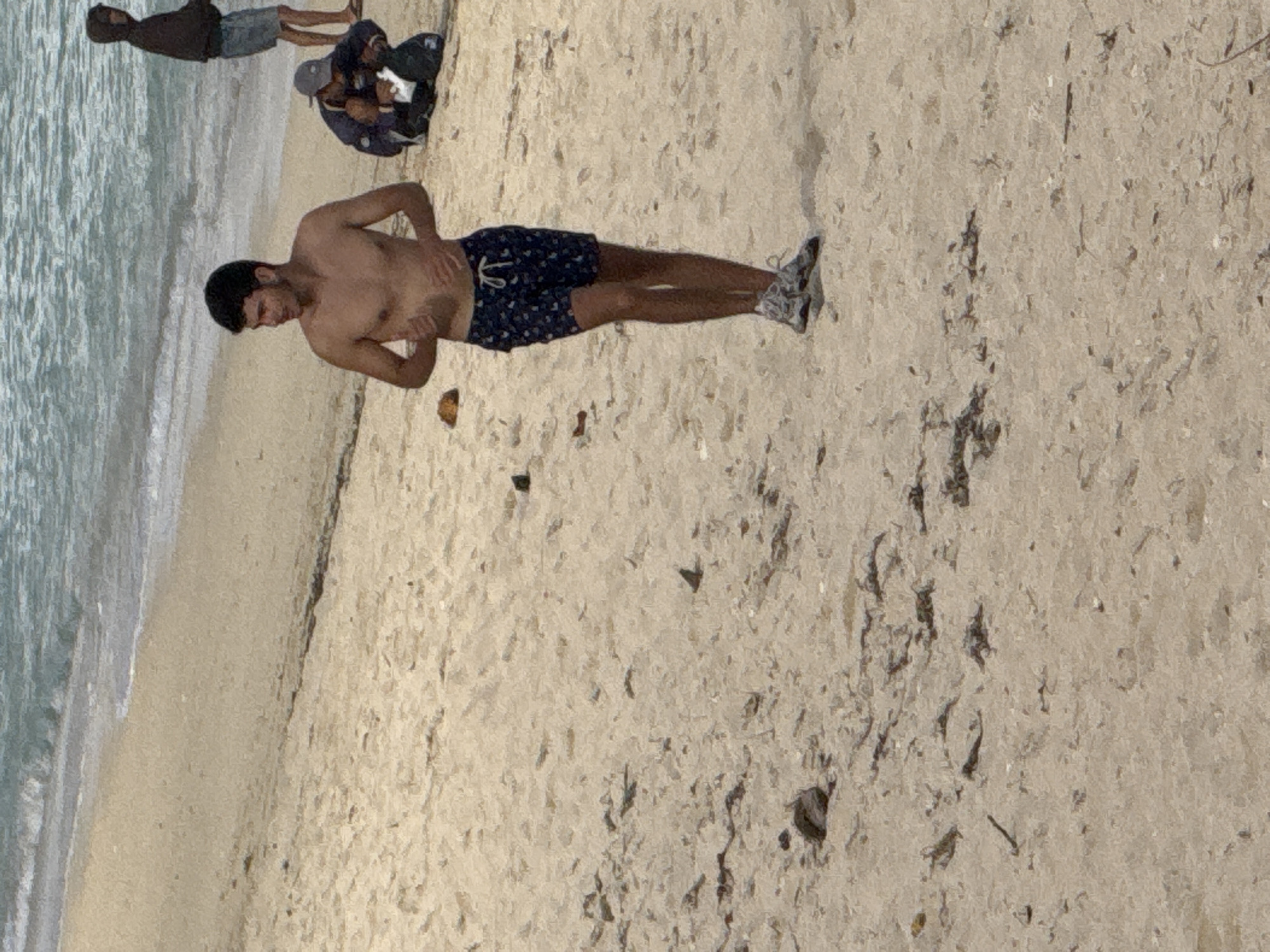 The image shows a sandy beach with the ocean in the background. Three people are visible. In the foreground, a young man is lying on the sand, shirtless and wearing blue patterned swim shorts and white sneakers. He is looking down and has his hands on his stomach. Behind him, two other individuals are partially visible, sitting on the sand with their backs to the camera. One appears to be wearing a dark hoodie, and the other has on a blue top and a grey baseball cap. The scene suggests a casual day at the beach. The lighting indicates it is daytime, and the water and sand suggest a tropical or coastal environment, consistent with the provided location context of Kuta Selatan, Indonesia. There is no visible text in the image.