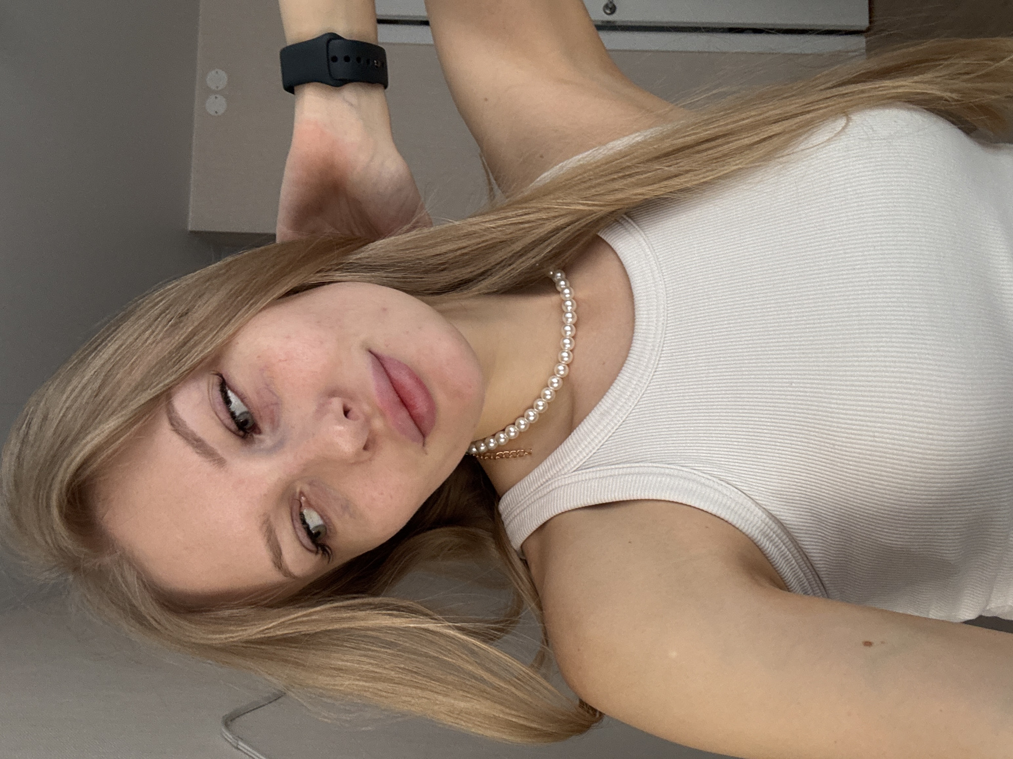 A young, fair-skinned woman with long blonde hair is seen close-up, wearing a white ribbed tank top and a pearl necklace with a gold pendant. A black smartwatch is visible on her raised wrist. The background is a neutral gray wall. The location is unspecified, and there is no indication it is in Perm, Russia.