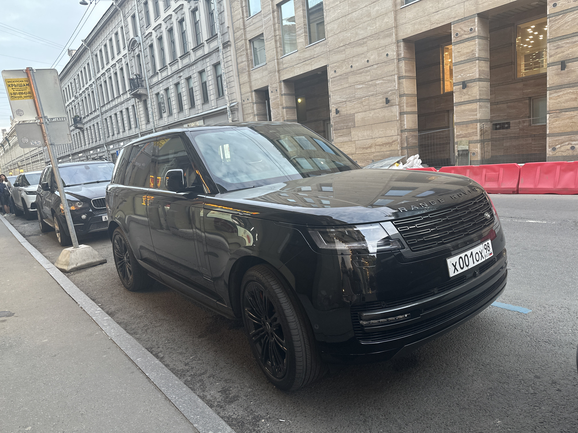 The image shows a black Range Rover SUV parked on a city street in Saint Petersburg, Russia. The license plate reads "X001OX 98." The car is parked next to a gray curb and a street sign with Cyrillic script. In the background, there are several light-colored buildings with classical architectural details on the left and a more modern building with light brown stone on the right. Other vehicles are parked along the street as well. There are red plastic barriers visible near the modern building.