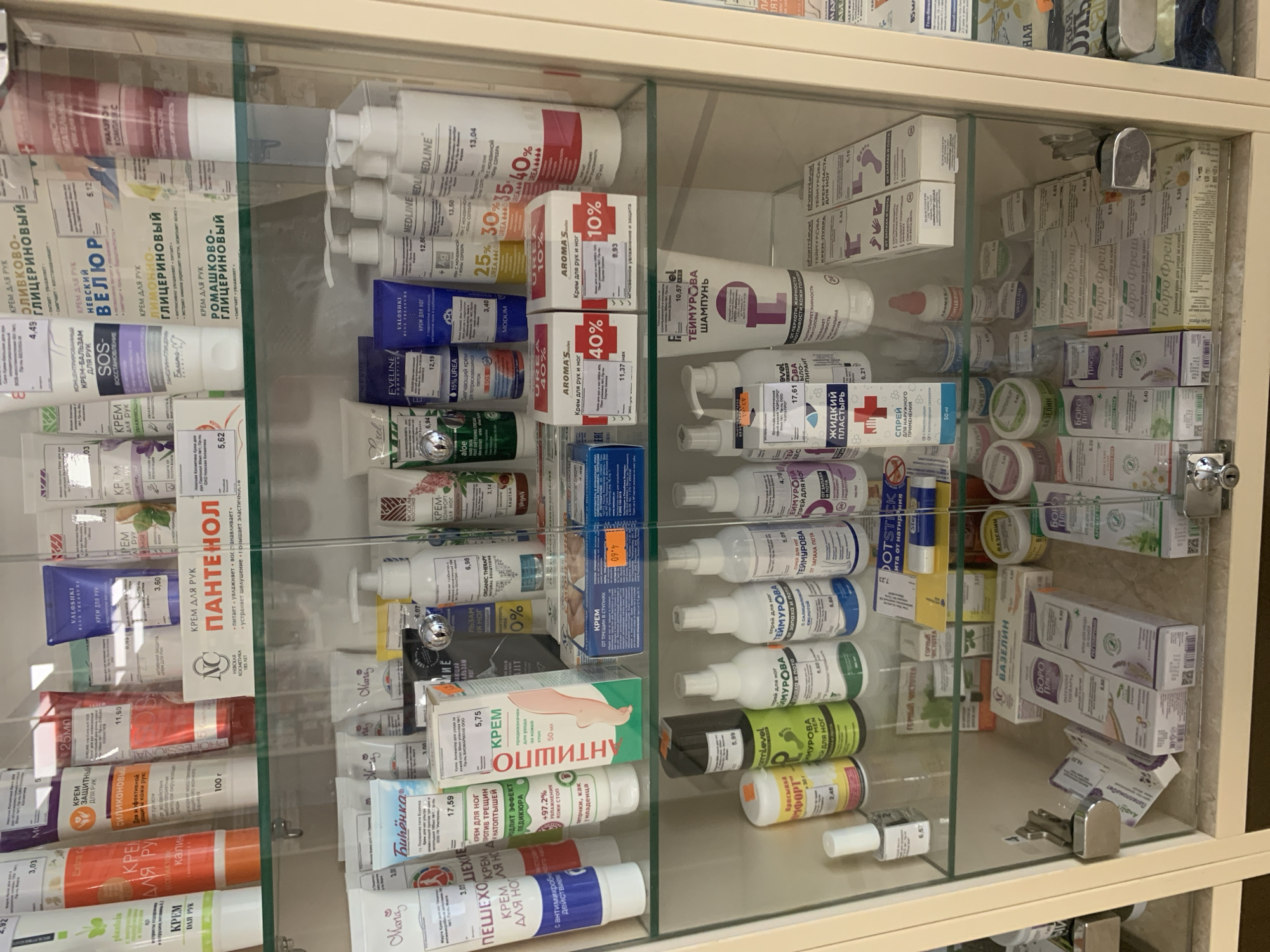 This is a retail display shelf in a pharmacy in Kapyĺ, Belarus. The shelves are stocked with a variety of personal care products, primarily creams, lotions, and sprays for skin and foot care. The products are in tubes, bottles, and boxes, with prominent branding and text in Russian. The overall environment is well-lit, suggesting an indoor commercial setting. No people are visible in the image. The display is organized with products arranged by brand and type.