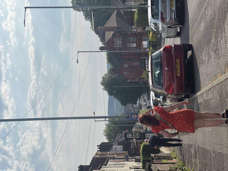An image of a street scene shows a red-haired woman in an orange dress standing on the sidewalk next to parked cars. Behind her are buildings and a street sign showing a speed limit of 30. The sky is light blue with clouds. In the background, there are distant hills. A man in dark clothing is walking away from the camera, closer to the building. Streetlights line the road. The scene appears to be a typical residential street on a sunny day.
