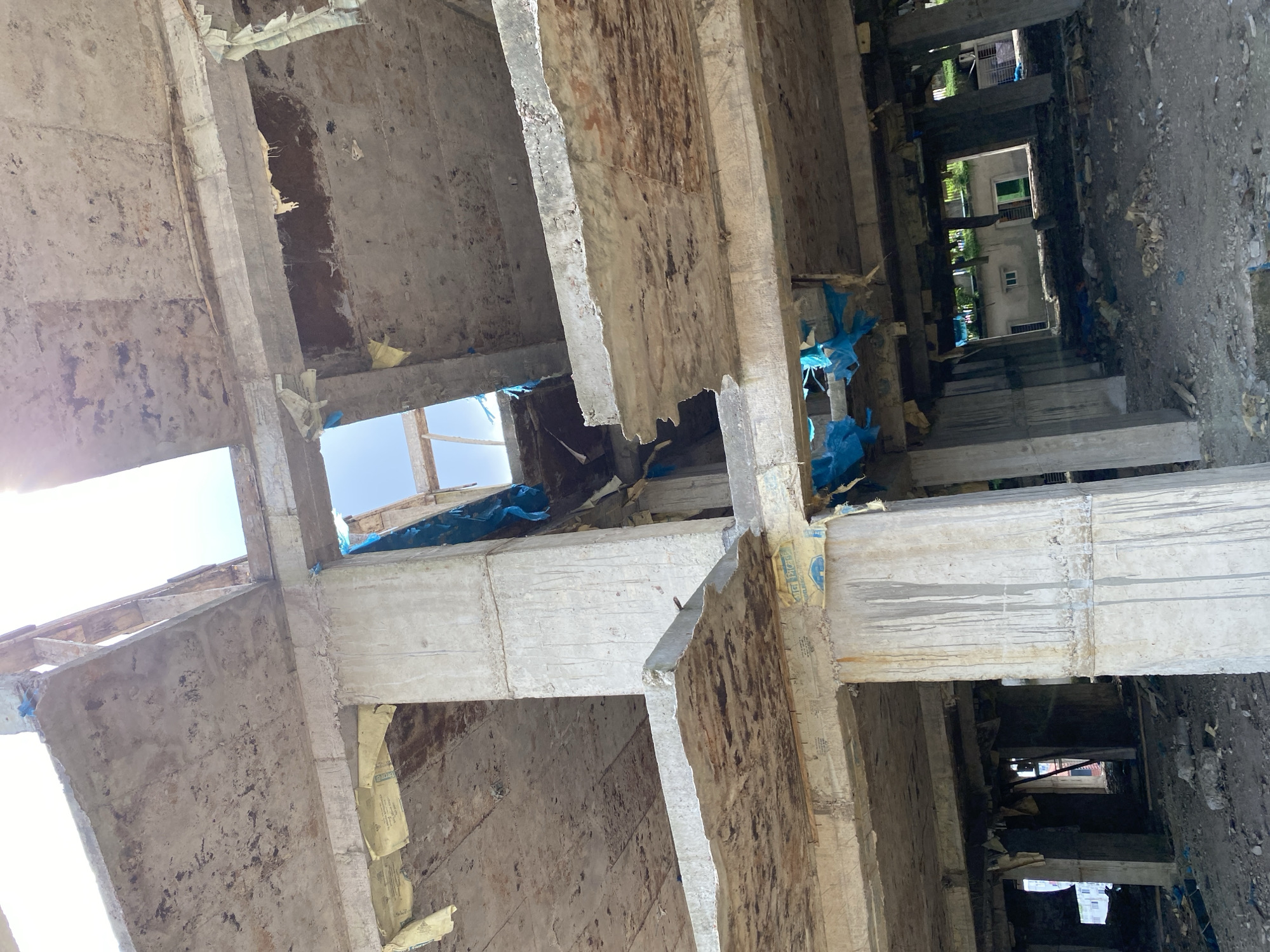 The image depicts the partially demolished interior of a multi-story concrete structure, likely under construction or renovation, in Dhaka, Bangladesh. The scene is characterized by exposed concrete beams and slabs, many of which show signs of damage and deterioration. Torn blue plastic sheeting is visible hanging from some of the structural elements, and yellowed paper or plastic sheeting is also present, possibly remnants of construction materials or protective coverings. The floors are littered with debris, including concrete fragments and other unspecified materials, indicating an ongoing or recently halted construction or demolition process. The sunlight streams through openings in the ceiling, illuminating the dusty environment. In the background, glimpses of other buildings and vegetation can be seen through the skeletal structure, suggesting an urban or semi-urban setting. No people are present in the immediate scene. The overall impression is one of decay and unfinished work.