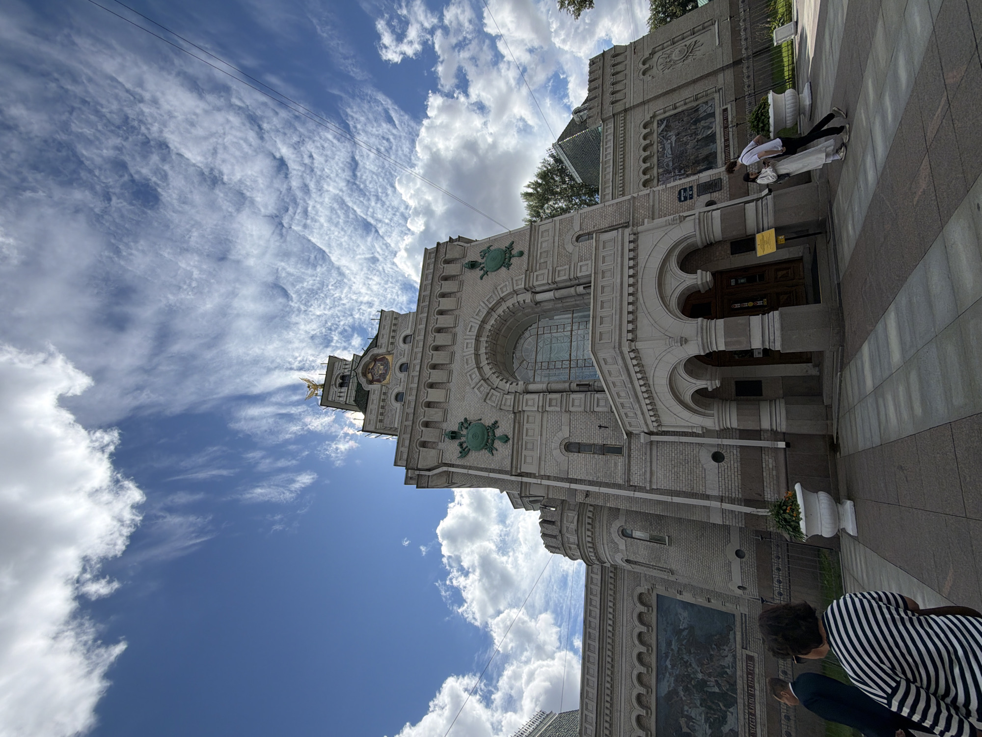 A low-angle, wide shot captures the neoclassical facade of the Naval Cathedral of Saint Nicholas in Kronstadt, Saint Petersburg, Russia, under a partly cloudy sky. The light-colored brick building features a central, arched entranceway with intricate stonework, flanked by two smaller arched entrances. Above the main entrance, a large, arched window is visible. The building's facade is adorned with decorative elements, including green circular crests and a mural depicting a battle scene on the left side. A clock is visible at the top of a tower, which is topped with a golden statue of an angel. Paved walkways with a checkered pattern lead up to the entrance. Two people are walking away from the camera on the left, while a couple stands near the entrance on the right, one person leaning against the other. Large white planters with orange flowers are placed in front of the building. Overhead power lines are visible against the sky.