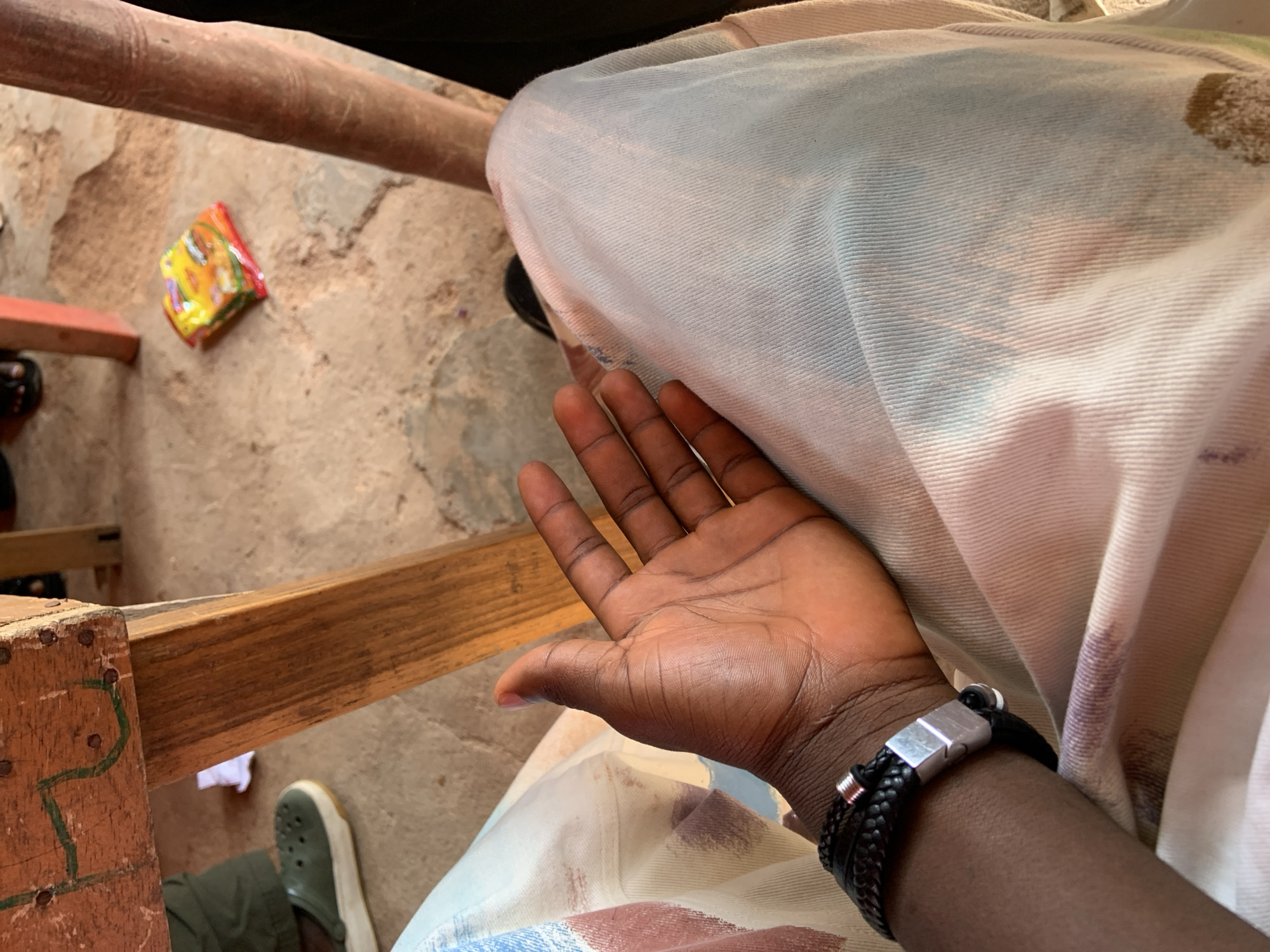 A close-up shot shows a person's left arm and hand resting on their lap in what appears to be an outdoor setting in the city of Jos, Nigeria. The person's skin tone is dark, and they are wearing a black braided bracelet with a metallic clasp on their wrist. Their hand is open, palm facing upwards, with fingers slightly curled. They are seated, with their legs covered by a light-colored fabric with a subtle pattern of pink and blue hues.
In the background, a portion of a wooden table leg is visible, its surface weathered and marked with what seems to be green paint or chalk drawings. To the left, the green rubber sole of a Croc-style shoe is peeking out from beneath the table. The ground is covered in sandy soil, with small pebbles and debris scattered about. A bright yellow and red packet of instant noodles or snacks lies on the ground behind the table leg. Another reddish wooden leg of what could be a chair or table is also visible in the background. The overall lighting suggests daylight, possibly indicating a casual or informal gathering.