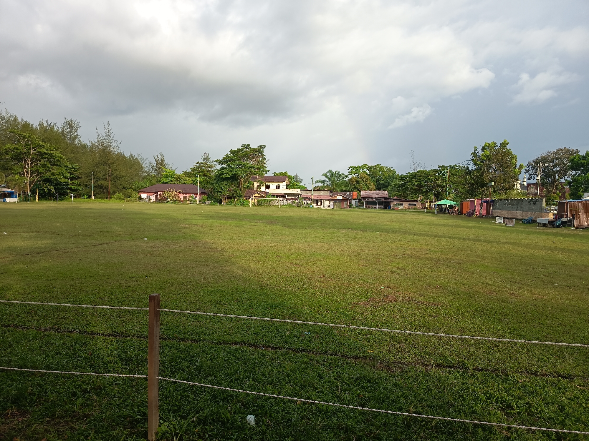 A wide, grassy field, likely a sports ground, occupies the foreground and midground of the image, sloping gently upwards towards the background. A simple rope fence with wooden posts runs across the bottom of the frame, separating the viewer's vantage point from the field. The field itself is a vibrant green, with some darker patches and lighter sunlit areas indicating unevenness or varying grass growth. In the midground, along the tree line that borders the field, are several buildings, some appearing to be residential houses with reddish-brown roofs, while others might be community structures or shops. A white soccer goal is visible on the left side of the field, near the tree line. The background is dominated by a dramatic, cloudy sky with patches of sunlight breaking through, suggesting an overcast or changing weather day. Various trees, including palm trees and taller pines, are interspersed with the buildings and form a dense line along the edge of the field, creating a natural backdrop. On the right side of the field, further back, are some makeshift stalls or covered structures, and beyond that, more buildings and trees. The overall impression is of an outdoor public space in a tropical or sub-tropical environment, possibly a community park or sports facility in Singkil, Indonesia, given the vegetation and architectural styles. No people are visible in the frame.