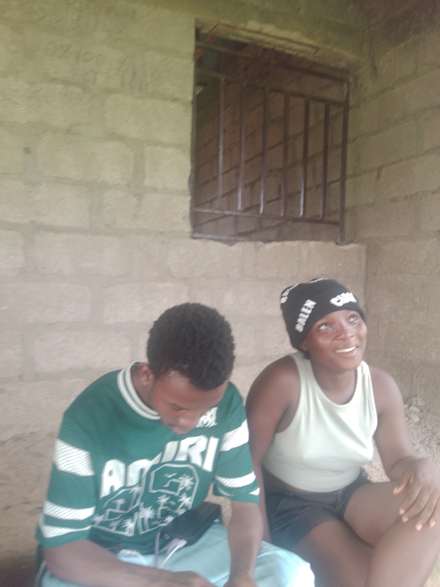 Two young individuals are seated outdoors in front of a concrete block wall. The person on the left, wearing a green and white striped jersey with the word "AMIRI" across the front and a graphic of palm trees, is looking down at a black device resting on their lap, which has wires plugged into it. The person on the right, wearing a light green tank top and black shorts, has a black beanie on their head with white lettering that reads "BALEN." This individual is looking upwards and smiling. Behind them, a window covered with metal bars is visible in the concrete wall, and the ground is sandy. The scene appears to be in Jauro Yinu, Nigeria.