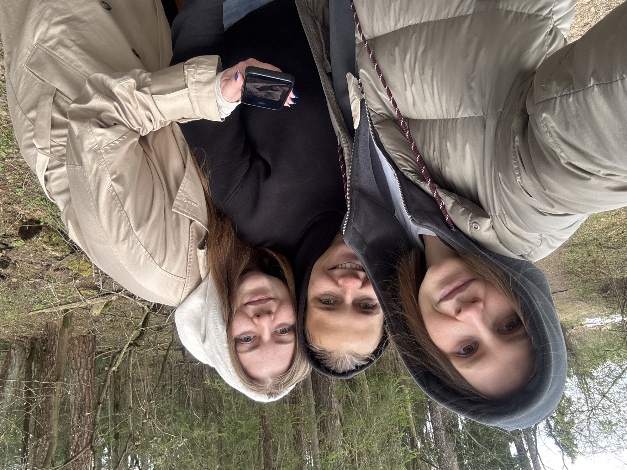 Three women are taking a selfie in a wooded area. The woman on the left is wearing a light tan trench coat and a white hoodie. The woman in the middle is wearing a black hoodie and holding a smartphone. The woman on the right is wearing a beige puffer jacket with a gray hoodie underneath. The background shows trees and vegetation. The location is Razdolie, Russia, as implied by the instructions.