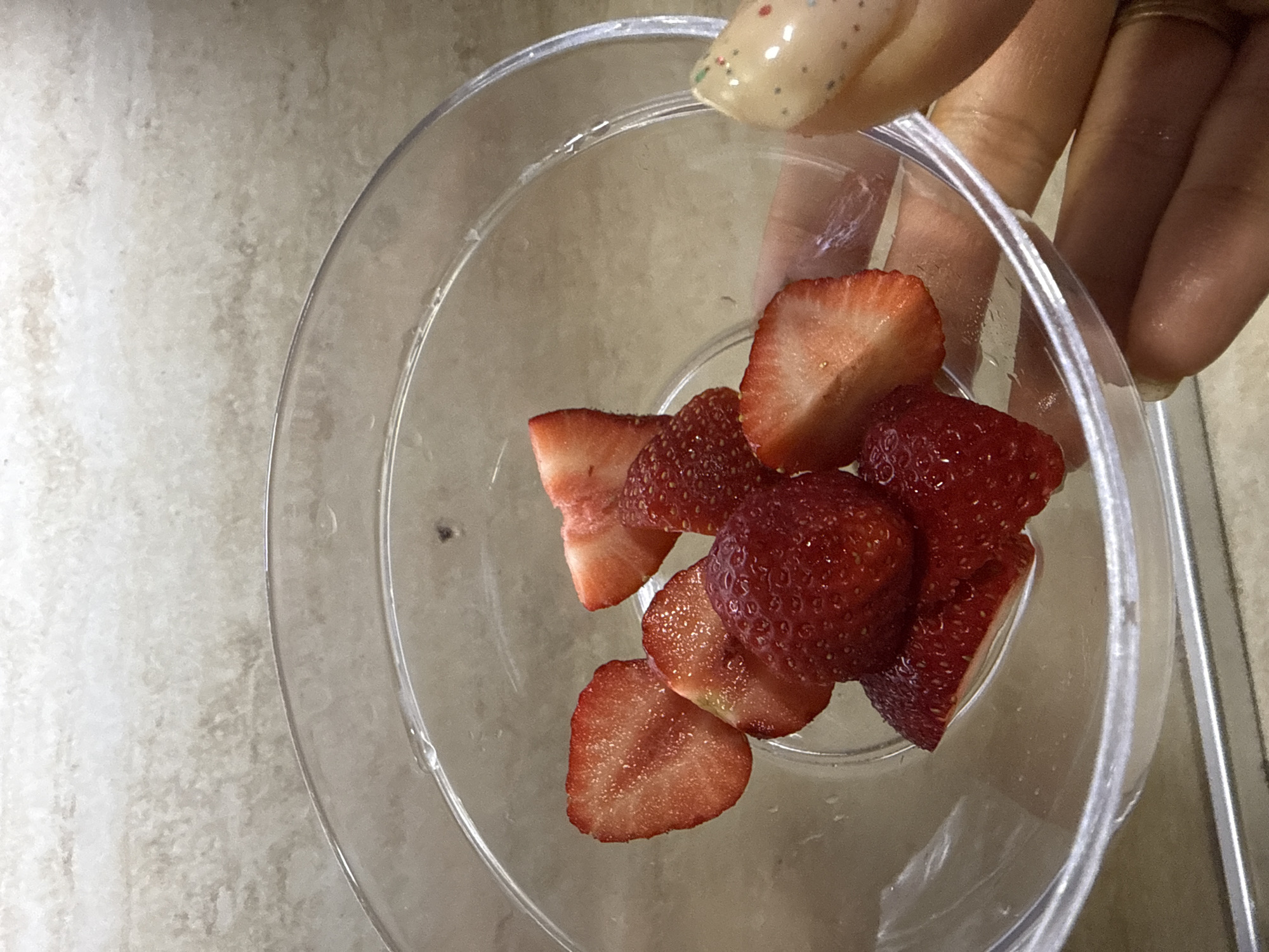 A close-up, high-angle shot shows a clear plastic container held by a hand with painted nails. The container holds approximately seven sliced strawberries. The background appears to be a light-colored countertop or surface. There is no immediate evidence to connect the image to Troyan, Bulgaria.