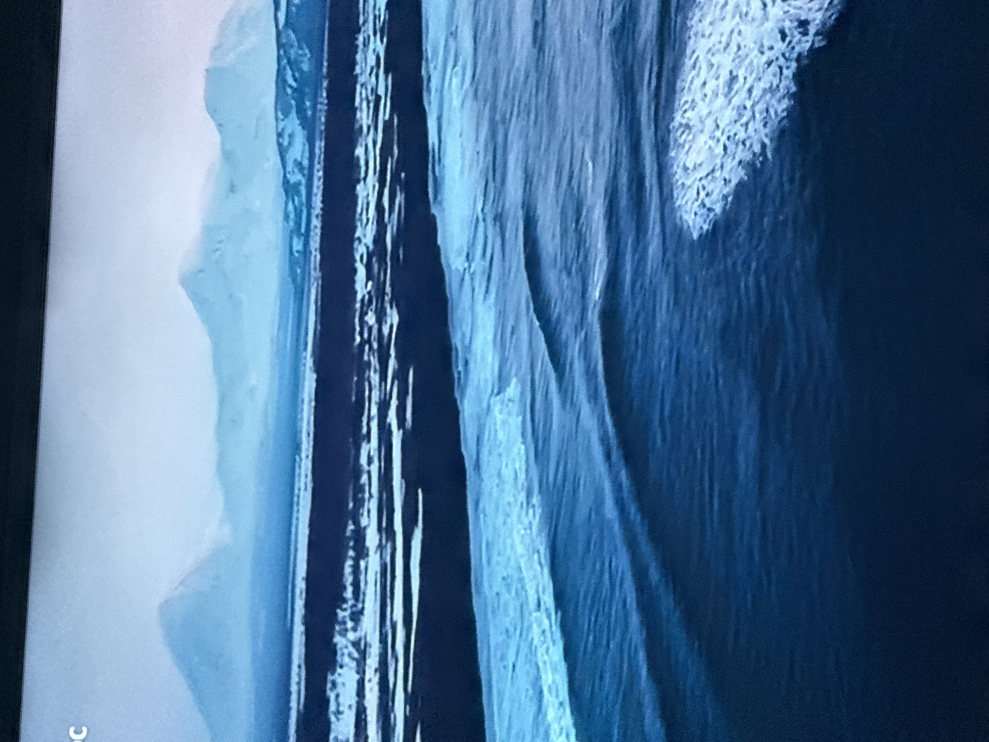 The image shows a vertically oriented close-up of a screen displaying a landscape, likely of icebergs or glacial formations. The palette is dominated by varying shades of blue and white. On the left, we see a distant, hazy mountainous region with a pale, almost cloud-like appearance. Progressing rightward, a narrow strip of what seems to be water or ice reflects the sky. Adjacent to this strip is a section of towering, vertically oriented ice or rock formations, streaked with meltwater or ice patterns. Further to the right, a substantial section of the image features the rugged, detailed texture of an iceberg, potentially with frosted or snowy features. The lower left corner of the image has the letters "C". The image does not contain people. The location is not Mytishchi, Russia.