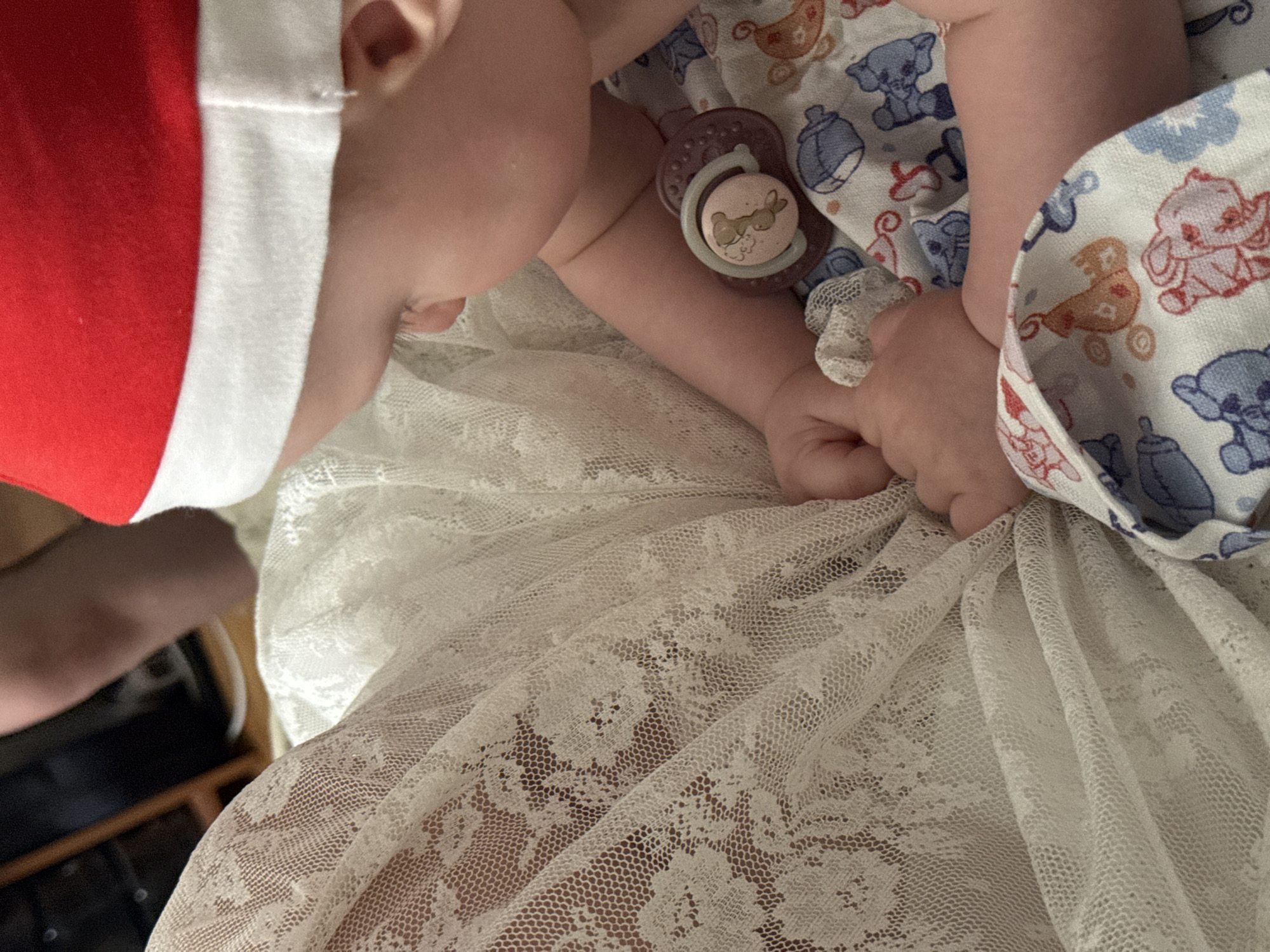 A close-up, overhead shot focuses on a baby lying down, covered partially by a white lace fabric. The baby is wearing a red and white Santa hat, suggesting a holiday or festive theme. A pacifier with a rabbit design is visible in the baby's mouth. The baby is also wrapped in a patterned fabric featuring illustrations of baby animals and objects like bottles and carriages. The background includes a glimpse of what appears to be an adult's leg and a dark surface, possibly furniture. The lighting suggests an indoor setting, and the overall mood is one of peacefulness and comfort. The image does not contain any visible text. Given the context of Moscow, Russia, this image might have been taken during the winter holiday season.