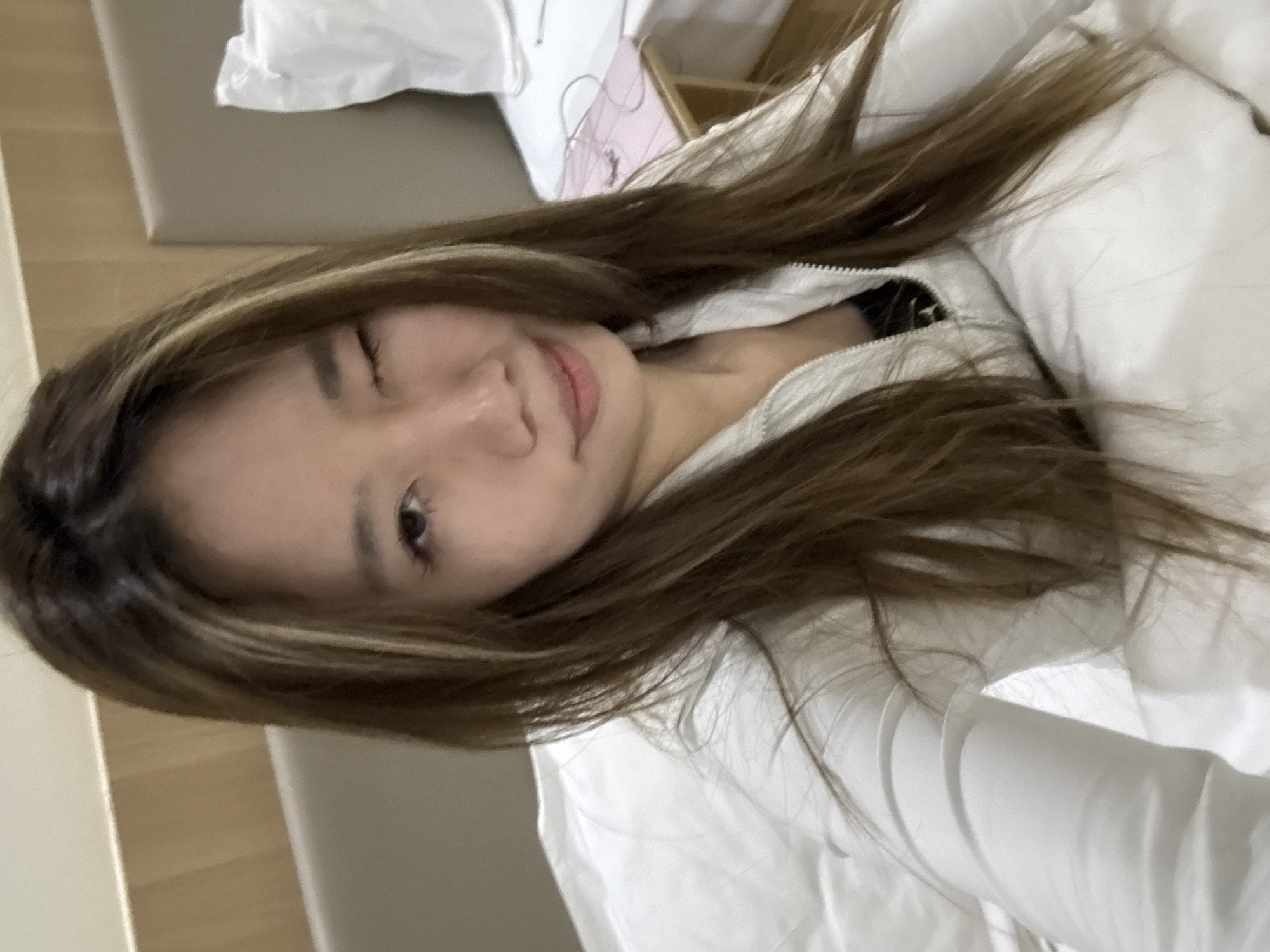 A medium, rotated indoor shot depicts a woman lying on a white bed. She is turned towards the camera, winking her left eye. Her hair is long, brown with blonde highlights, and slightly disheveled. She appears to be wearing a white jacket or top. The background is blurry but includes a beige surface, possibly part of the bed frame or a side table, along with white bedding. No information in the photo indicates the location as Dunhuang City, China.