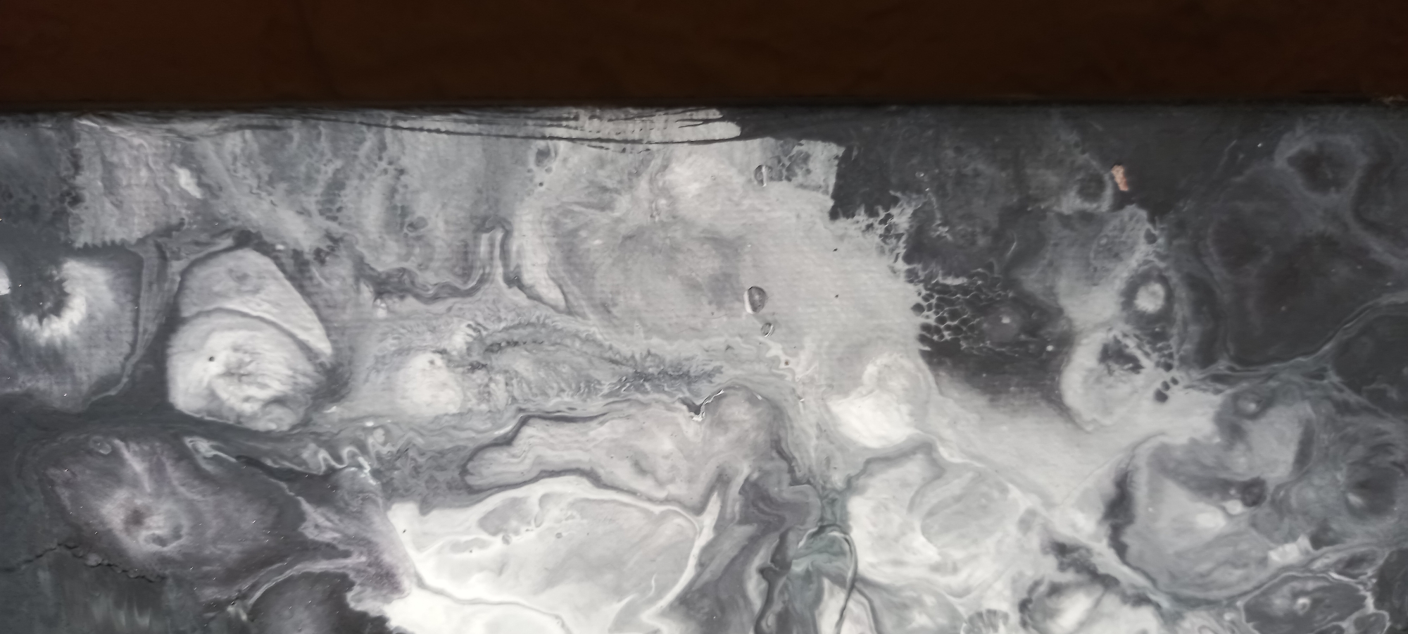 The media file presents a close-up view of an abstract artwork, primarily composed of black, white, and various shades of grey. The painting exhibits characteristics of fluid art, such as acrylic pouring, with intricate swirling, marbling, and cellular patterns throughout its surface. The organic forms create a sense of movement and depth, resembling natural elements like clouds, smoke, or liquid flows.
The artwork fills most of the frame, with a dark brown, possibly wooden, surface visible along the top edge, suggesting it might be resting against or on an adjacent object. The surface of the painting appears to have a slight gloss or sheen. Specific details include several small, dark specks visible in various areas of the fluid patterns, and a small, reddish-brown imperfection or inclusion located within a darker grey section towards the upper right quadrant of the image.
There are no people, identifiable specific objects beyond the artwork itself, or activities taking place. The setting appears to be indoors, indicated by the plain background beyond the artwork. No time of day, weather conditions, or specific location cues are discernible from the abstract content of the image. There is no visible text.