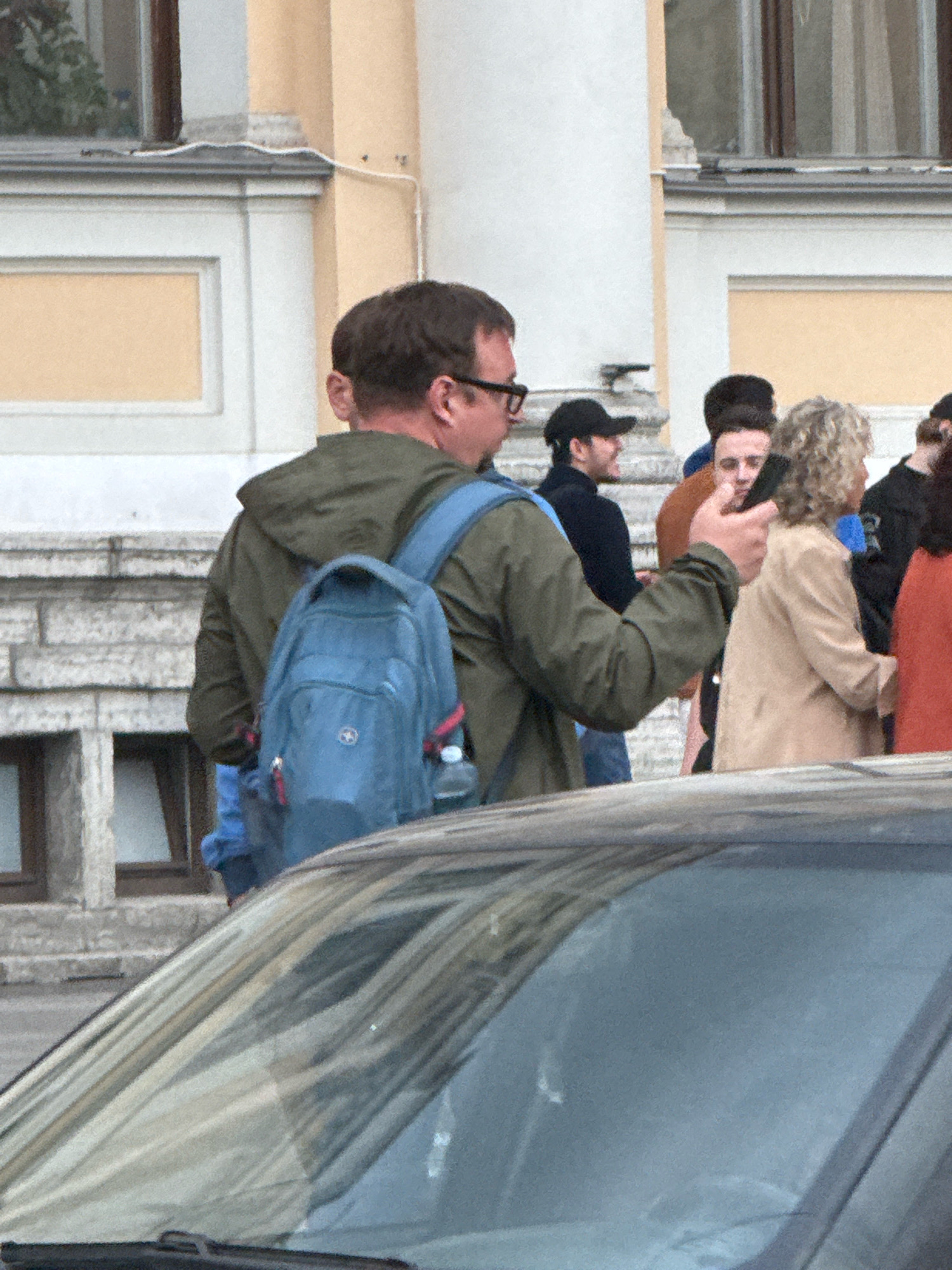 A group of people are gathered outdoors, possibly in front of a building with classical architectural elements, suggesting a location in Saint Petersburg, Russia, such as near a historical landmark or cultural institution. The foreground shows the hood and windshield of a dark-colored car, partially obscuring the lower half of the scene.
In the mid-ground, a man wearing glasses and a green jacket with a blue backpack is the primary subject. He is turned away from the camera, looking towards the right. A blue backpack with red accents is visible on his back. To his right, several other individuals are present. One person, wearing a light-colored jacket, is holding a mobile phone. Other people in the background are less distinct but appear to be in conversation or observing their surroundings. The building in the background features large windows and decorative stonework. The lighting suggests it is daytime, possibly overcast.