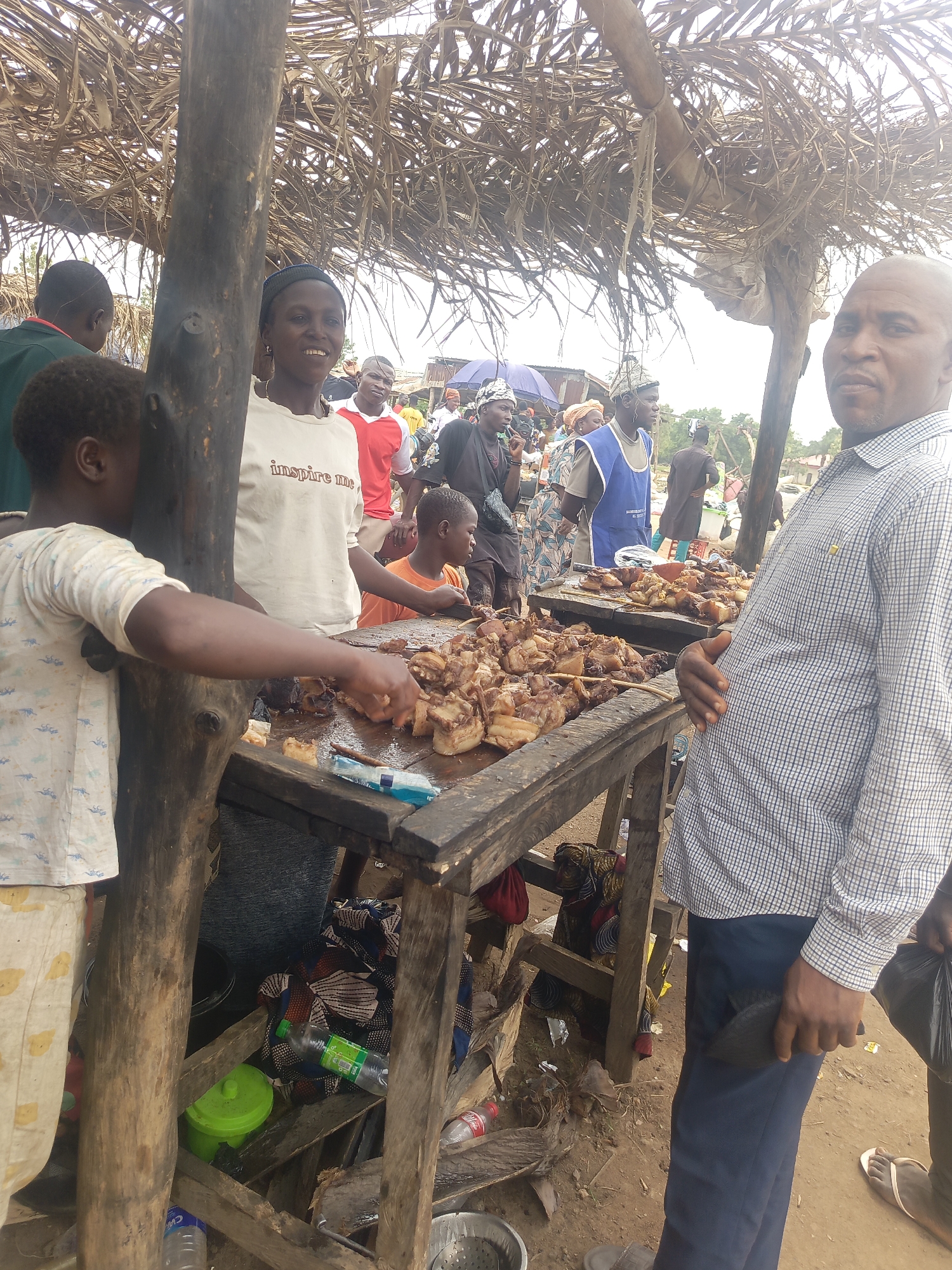 This media file captures a vibrant outdoor market scene in Andaha, Nigeria. The central focus is on two rustic wooden stalls laden with numerous skewers of cooked meat, likely grilled or roasted, which are being sold.
Several people are visible in the frame, indicating a busy atmosphere. On the left, a young child in a light-colored shirt is reaching towards the meat on one of the stalls. Next to the child, a woman wearing a white t-shirt with the text "inspire me" printed on it is smiling broadly towards the camera. Behind her, other individuals are partially visible, including a man in a red shirt.
To the right, a man in a blue and white checkered shirt is standing, holding a black bag, and looking directly at the camera. His posture suggests he might be observing the scene or posing. Further back, more people are gathered around other market stalls, some wearing traditional attire, under a canopy made of dried palm fronds or branches that provides shade.
Notable objects include plastic bottles of water and soda (a green bottle with "BIG" and a red Coca-Cola bottle are visible on the ground and under the stalls), and a green plastic container. The ground is unpaved earth. The overall lighting suggests it is daytime, with a bright, possibly overcast sky visible through the canopy. The scene depicts an active, open-air food market.