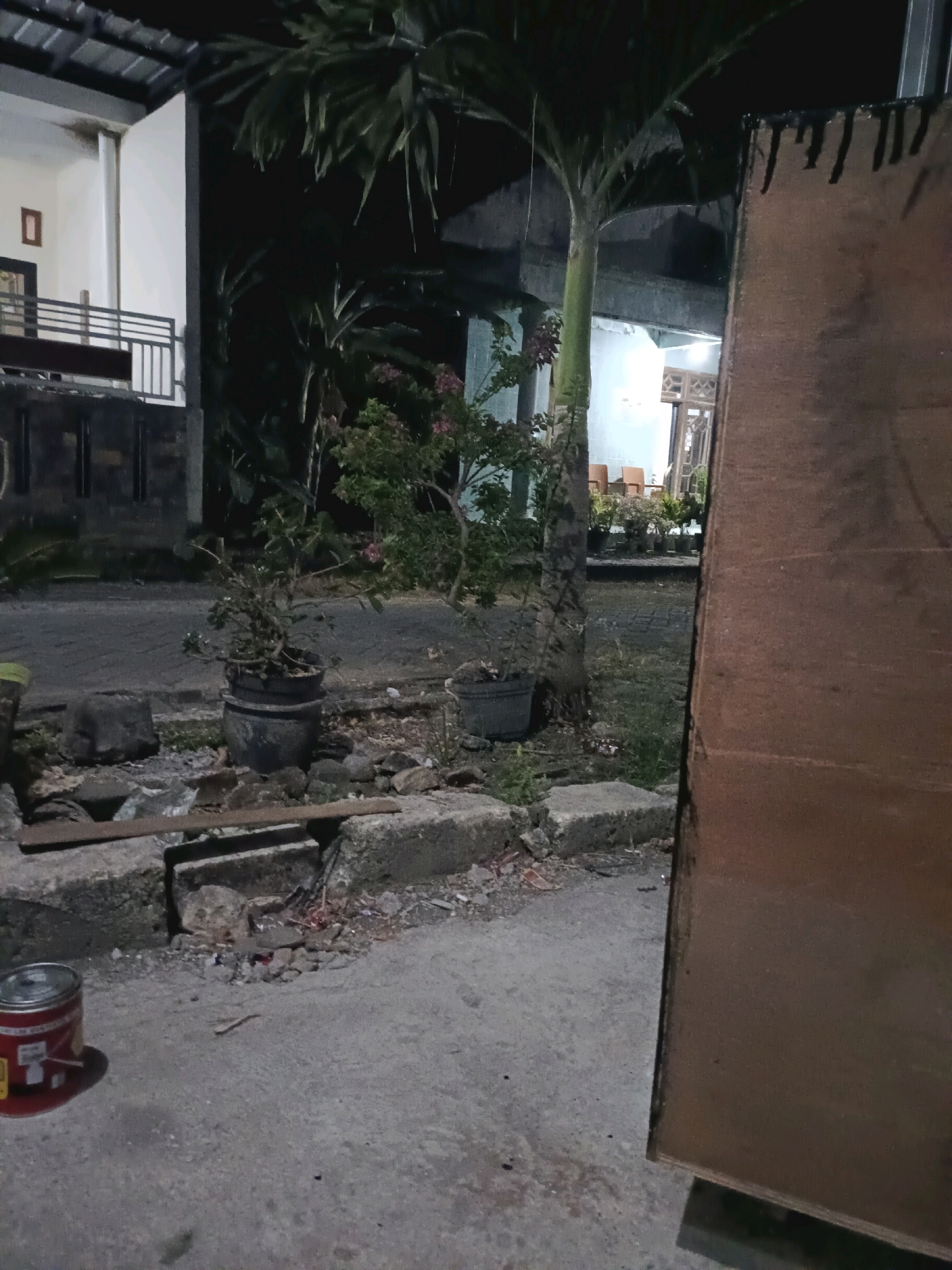 The image depicts an outdoor scene at night, likely in the city of Rancang Kencono, Indonesia. In the foreground, a red and silver can sits on a gray concrete surface. To the left of the can, there are several large, rough-hewn stones. Behind the stones, a potted plant with dark leaves is visible. Further into the background, a paved road runs horizontally. On the left side of the road, a white building with a dark-tiled roof and a balcony with a black railing is present. A small framed object hangs on the white wall of the building. A tall palm tree stands prominently in the center of the image, its fronds reaching upwards. To the right of the palm tree, another potted plant with green foliage and pink flowers is situated. Beyond this, a wooden structure, possibly a large board or wall, occupies the right side of the frame. In the far background, parts of buildings with illuminated windows are visible, along with several empty chairs and more potted plants, suggesting an outdoor seating area. The overall environment appears to be a residential or communal outdoor space at night, with a mix of natural elements and man-made structures.