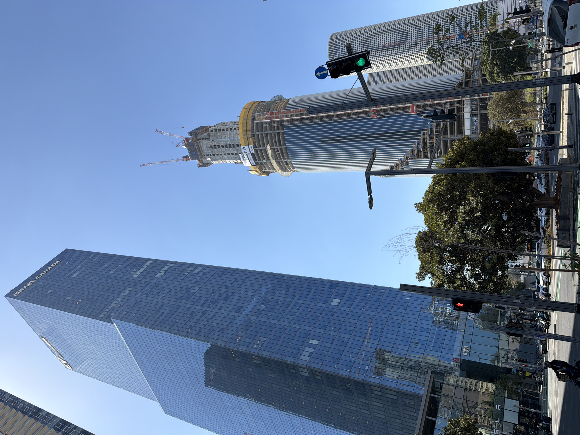 The image shows a street scene in Tel Aviv, Israel, with tall modern skyscrapers dominating the skyline. The "Israel Canada" building, a glass-clad skyscraper, is prominently featured on the left. In the center, another high-rise building appears to be under construction, with cranes visible on top. A third, cylindrical skyscraper is visible in the background. The foreground contains street elements such as traffic lights, some showing green and some showing red, trees, and sidewalks. There are cars and scooters parked along the street. People appear on the sidewalk, including a person on a bicycle. The sky is clear and blue.