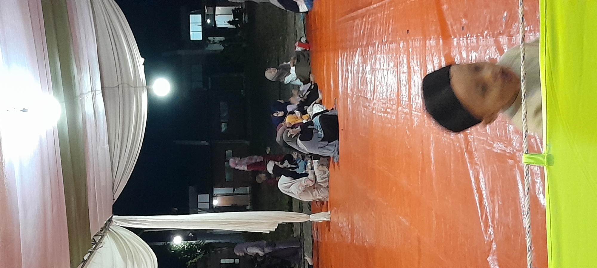 This image depicts an outdoor gathering at night, possibly a religious or social event, under a draped canopy. The setting appears to be in Bendungan, Indonesia, given the context provided. Several individuals, predominantly women wearing hijabs and modest clothing, are seated on an orange tarp. The lighting is provided by overhead lamps, casting a warm glow on the scene. In the foreground, a young child wearing a black cap is partially visible, looking towards the camera. The background shows dark silhouettes of buildings and trees, suggesting a semi-urban or suburban environment. The overall atmosphere is one of communal gathering and shared activity.