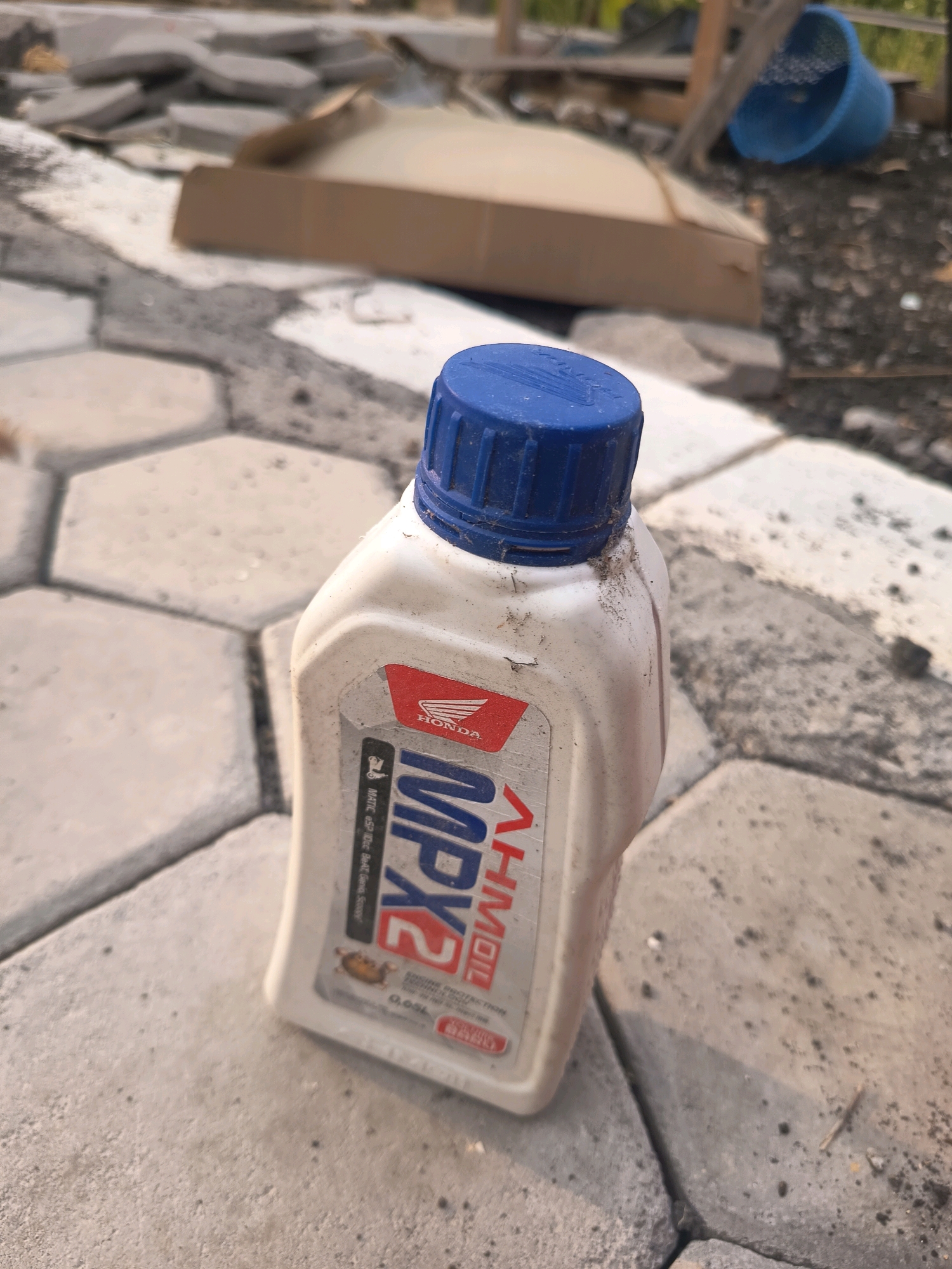 A dirty, white plastic bottle of Honda MPX2 motorcycle oil with a blue cap is lying on its side on a hexagonal patterned pavement. The bottle is positioned in the foreground of the image. The label on the bottle is partially visible and shows the Honda logo and "MPX2". In the background, there are scattered pieces of broken concrete, a cardboard box, and a blue plastic bucket. The environment appears to be an outdoor area, possibly a construction site or a yard, with a mix of rough ground and paving stones. The lighting suggests it is daytime, with shadows cast on the ground. The location is the city of Sidorejo, Indonesia.