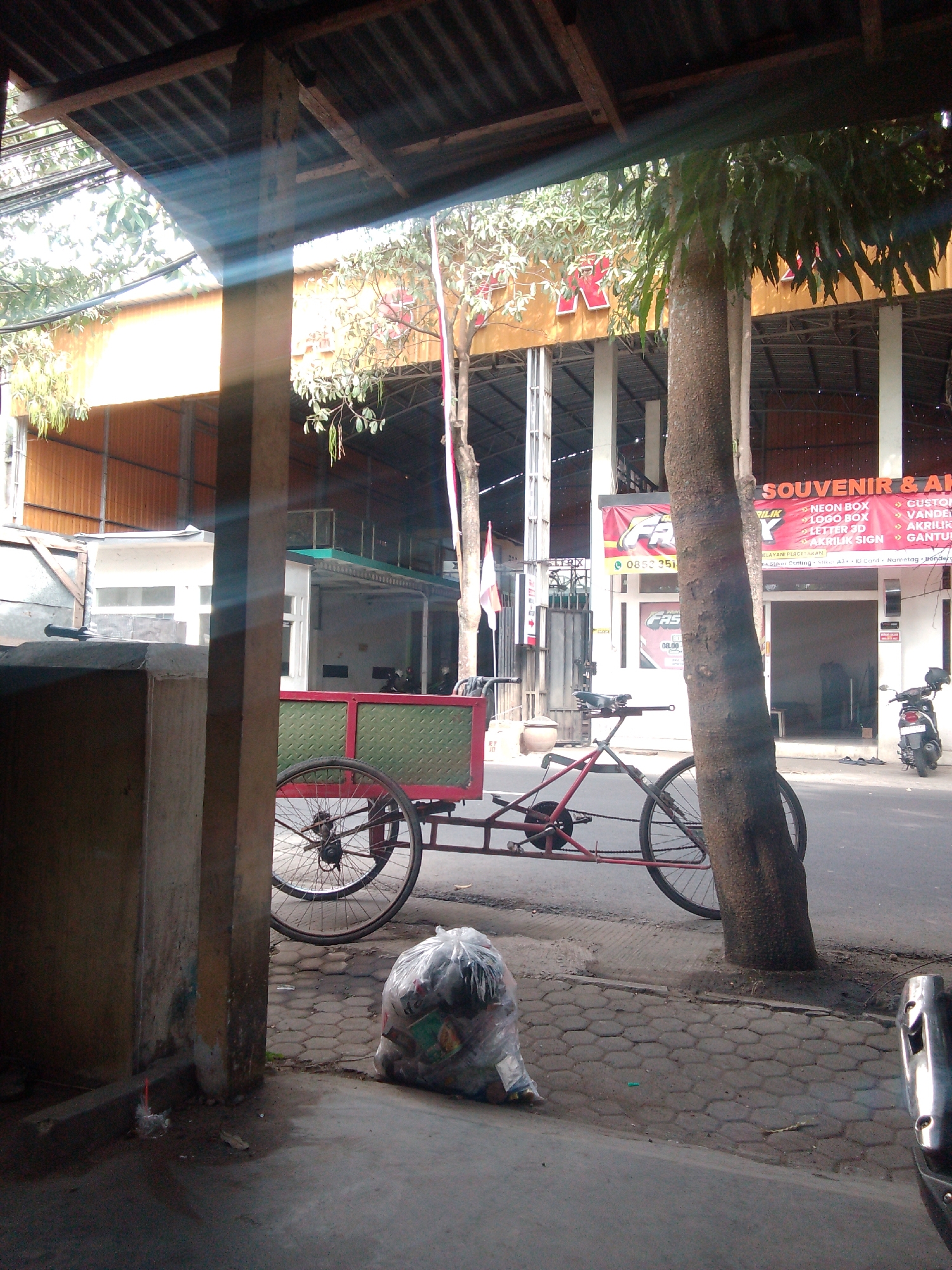 The image depicts an outdoor scene on a street in Trenggalek, Indonesia. In the foreground, partially obscured by a wooden structure, is a red and green cart attached to a bicycle. The cart is empty and has a patterned green side. The bicycle has two large spoked wheels, a red frame, and a black seat. To the right of the bicycle, a large tree trunk is prominently featured. A clear plastic bag filled with trash sits on the paved sidewalk to the left of the bicycle.
In the background, several buildings are visible. One building has a bright orange facade and a dark roof structure supported by white pillars. A banner with red lettering and the word "FAST" is displayed on this building, along with a phone number, "0852 3518". Another building, likely a shop, has a sign that reads "SOUVENIR & AK...". The street appears to be paved, and there are some trees and vegetation visible throughout the scene. A motorcycle is parked on the right side of the street. The lighting suggests it is daytime, with sunlight casting beams through the foliage and structures.
