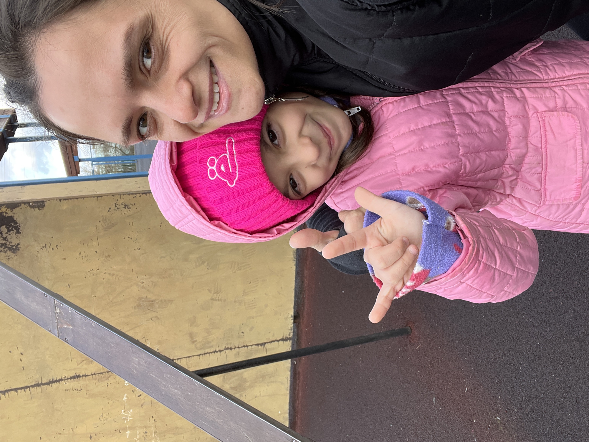 The image, rotated 90 degrees clockwise, captures a joyful selfie moment between an adult woman and a young girl, likely mother and daughter. Both subjects are smiling broadly, looking directly at the camera.
The woman, visible on the left side of the frame, has dark hair pulled back and warm brown eyes. She is wearing a black quilted jacket. To her right and slightly below, the young girl, also with dark hair and brown eyes, is dressed for cool weather in a vibrant bright pink quilted winter jacket. She wears a matching bright pink knitted beanie adorned with a distinctive white embroidered design that depicts the "I love you" sign in American Sign Language (thumb, index finger, and pinky extended). The girl's right hand is raised, making a similar gesture with extended thumb, index, and pinky fingers.
The setting is outdoors, likely on an overcast day, indicated by the diffused light and the warm attire worn by both individuals. The background features a textured, worn-looking wall or surface in shades of light brown and yellow. Below the subjects, a dark, possibly rubberized, ground surface is visible. A metallic or wooden beam or railing crosses the bottom left of the frame. A small portion of a cloudy sky and distant structural elements can be seen in the upper left background.
The overall mood is one of happiness and affection, conveyed through their wide smiles and direct gaze. The image portrays a candid, heartwarming interaction in an outdoor environment in Chaikovsky, Russia.