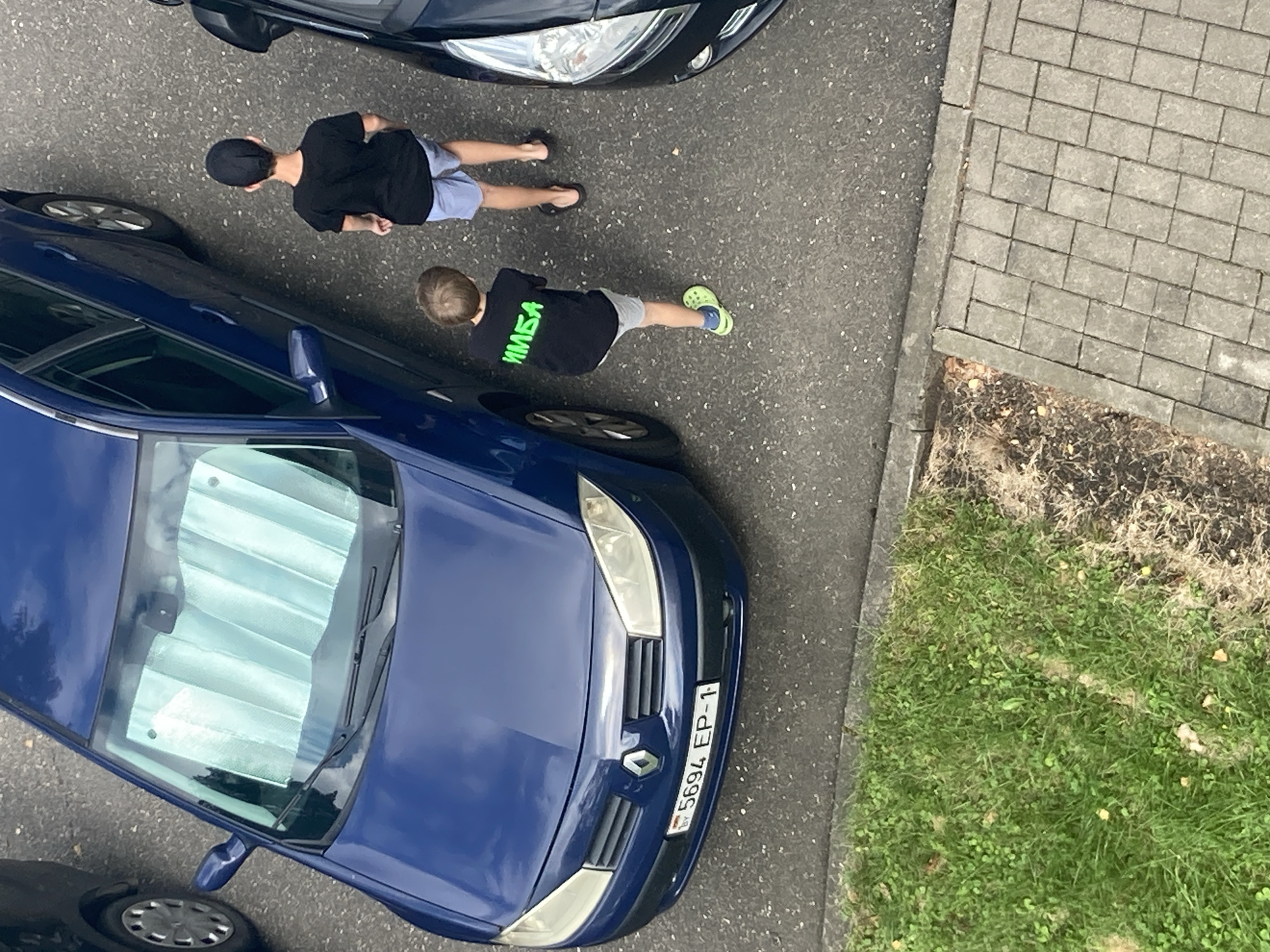 The image shows two young boys standing on asphalt next to a blue car. The boy on the left is wearing a black t-shirt with green lettering and grey shorts, and lime green crocs. The boy on the right is wearing a black t-shirt and light blue shorts, and black flip-flops. He is also wearing a black baseball cap. The blue car has a Belarusian license plate that reads "5694 EP-1". The scene appears to be in a residential area, possibly a parking lot, with grass and a paved walkway visible in the background. The image was taken from an elevated perspective, looking down at the scene.