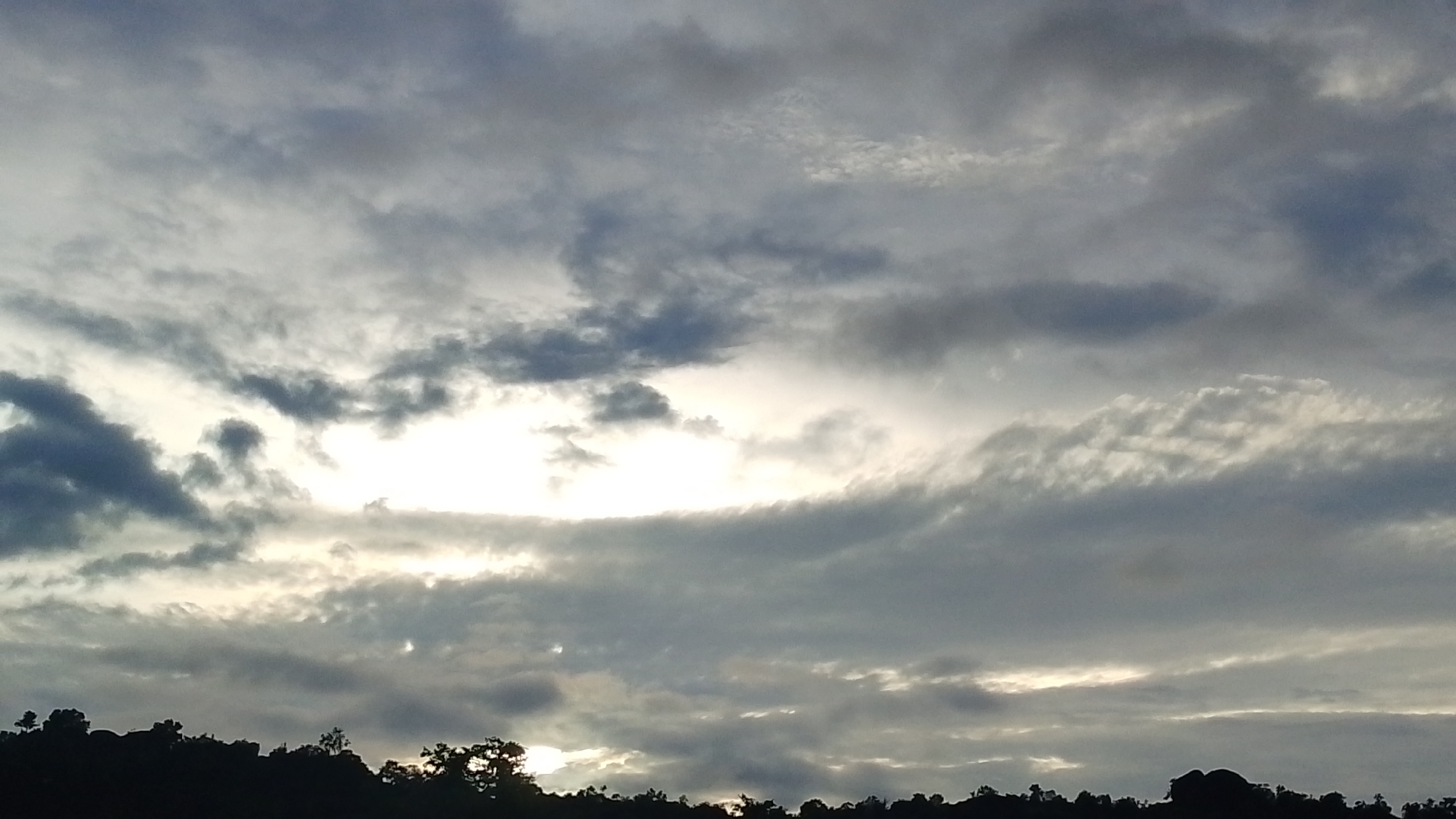 The image depicts a silhouetted landscape against a cloudy sky. The foreground shows the dark outlines of trees and possibly rocky outcrops along a ridgeline. Above this, the sky is filled with a dramatic arrangement of clouds, with patches of bright light breaking through, suggesting either sunrise or sunset. The lighting is diffused and atmospheric. No people or specific structures are discernible in the silhouette. The scene is likely outdoors and could be located in a natural or semi-natural area within the city of Indo, Nigeria, given the typical topography and vegetation found in such regions.